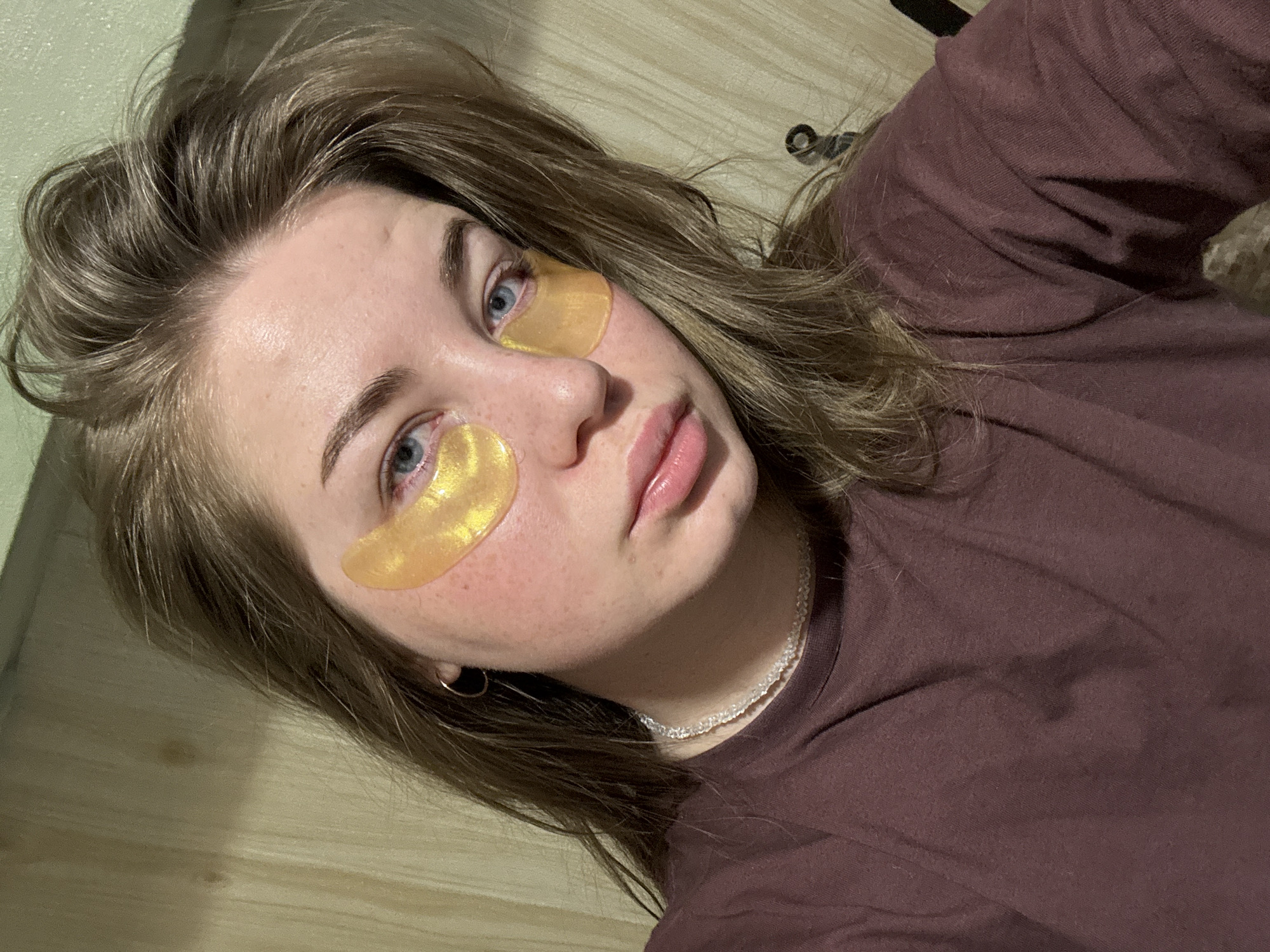 A young woman lies down, facing the camera. She has fair skin, blue eyes, and light brown hair. She has two gold under-eye patches applied. She wears a brown t-shirt and a white choker necklace. She is in an indoor setting with a light-colored wooden surface visible. There is no evidence to suggest the location is Chaikovsky, Russia.