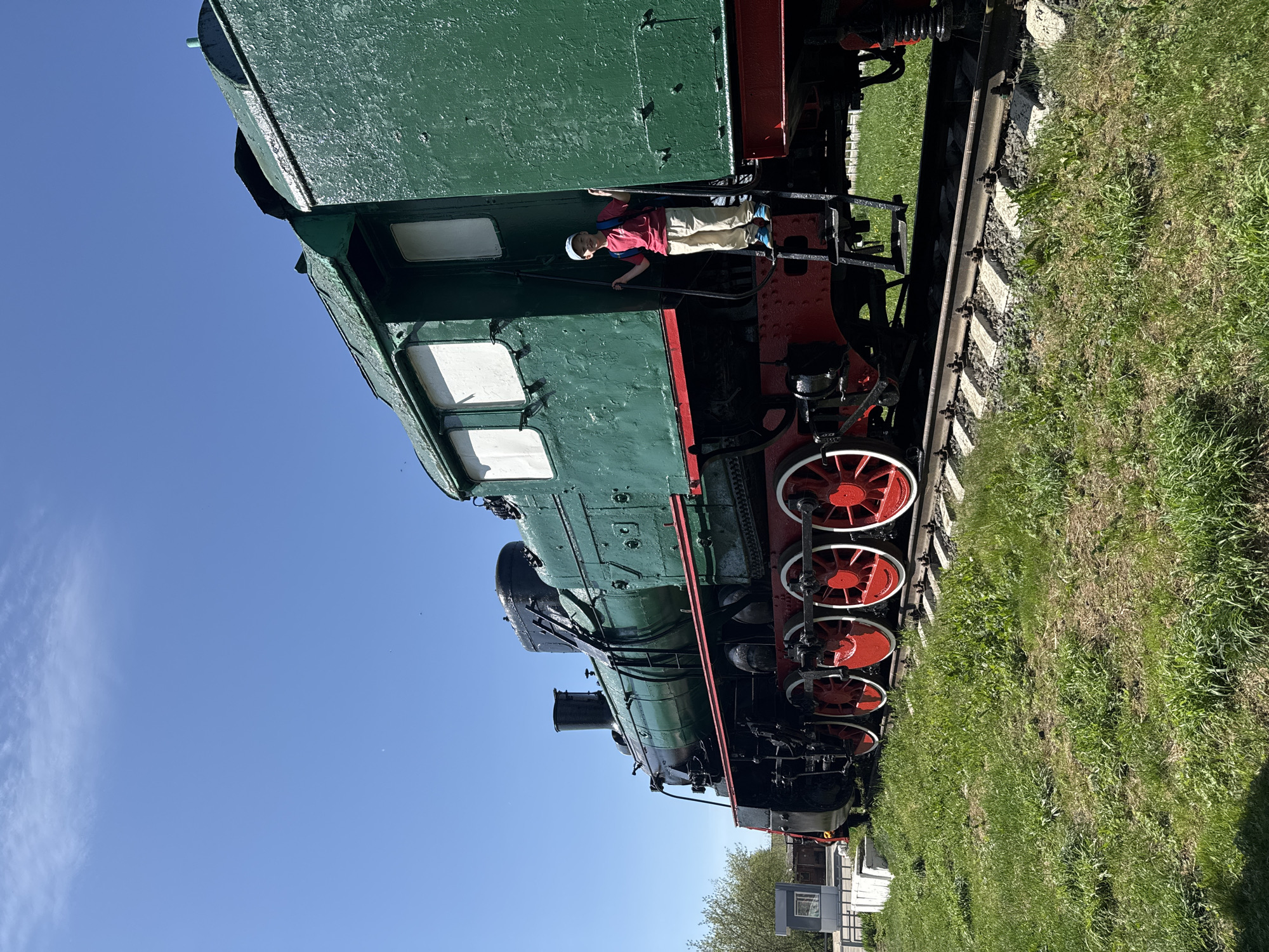 The image shows a green steam locomotive with red wheels, situated on a track in a grassy area under a clear blue sky. A young person wearing a red shirt, tan pants, and a light-colored cap is standing on the side of the locomotive. In the background, there is a small building and some trees. The location is likely Balezino, Russia, based on the prompt.
