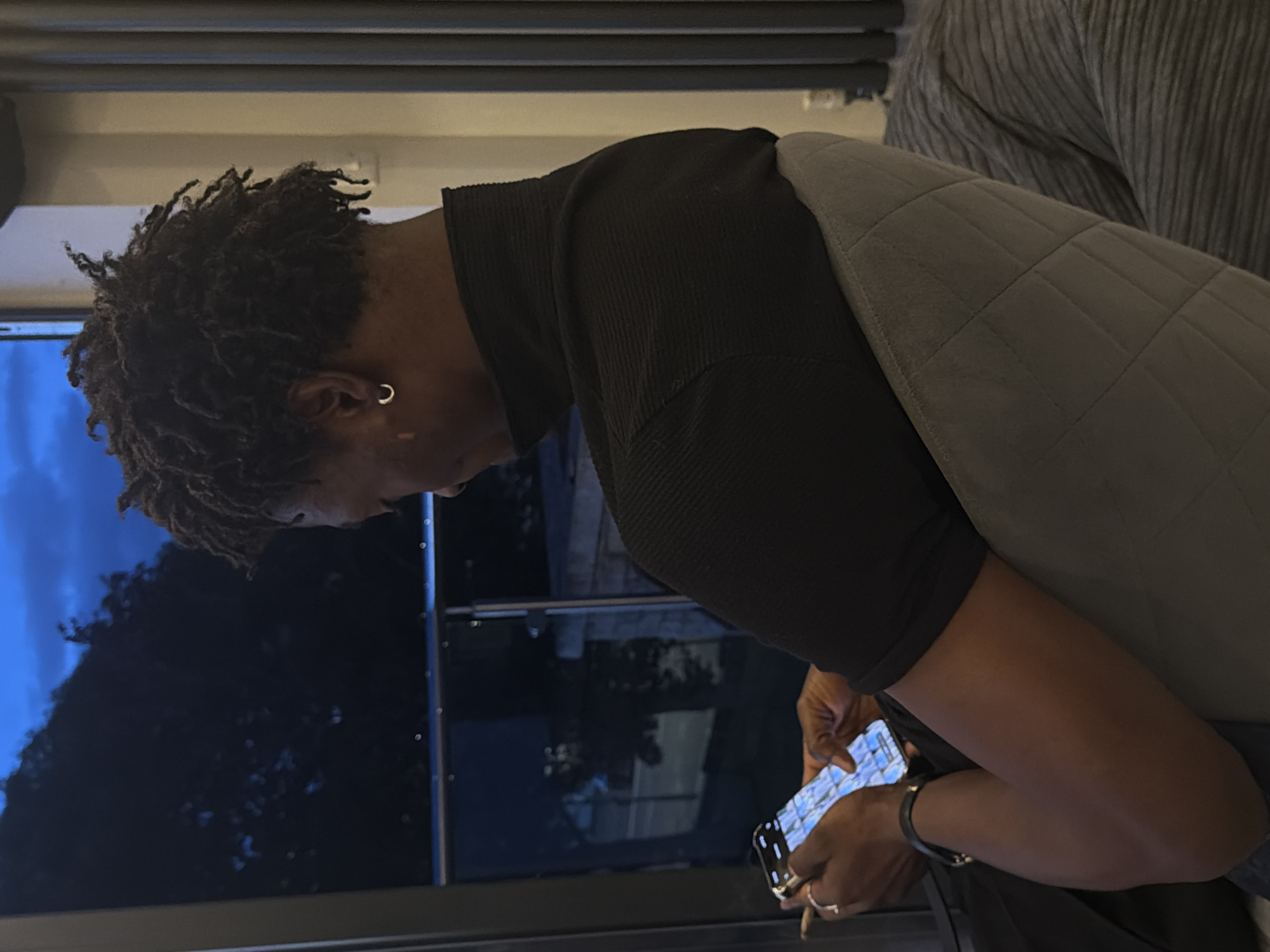 The image shows a person with dark skin and dreadlocks sitting in a chair, holding a smartphone. They are wearing a black turtleneck shirt and a ring on their left hand. The background appears to be a window overlooking a dark, tree-filled area with a blue sky. The person's posture and the way they are holding the phone suggest they are actively engaged with it. The lighting indicates it is evening or night. Without further context or identifiable landmarks, the specific location in London cannot be determined.