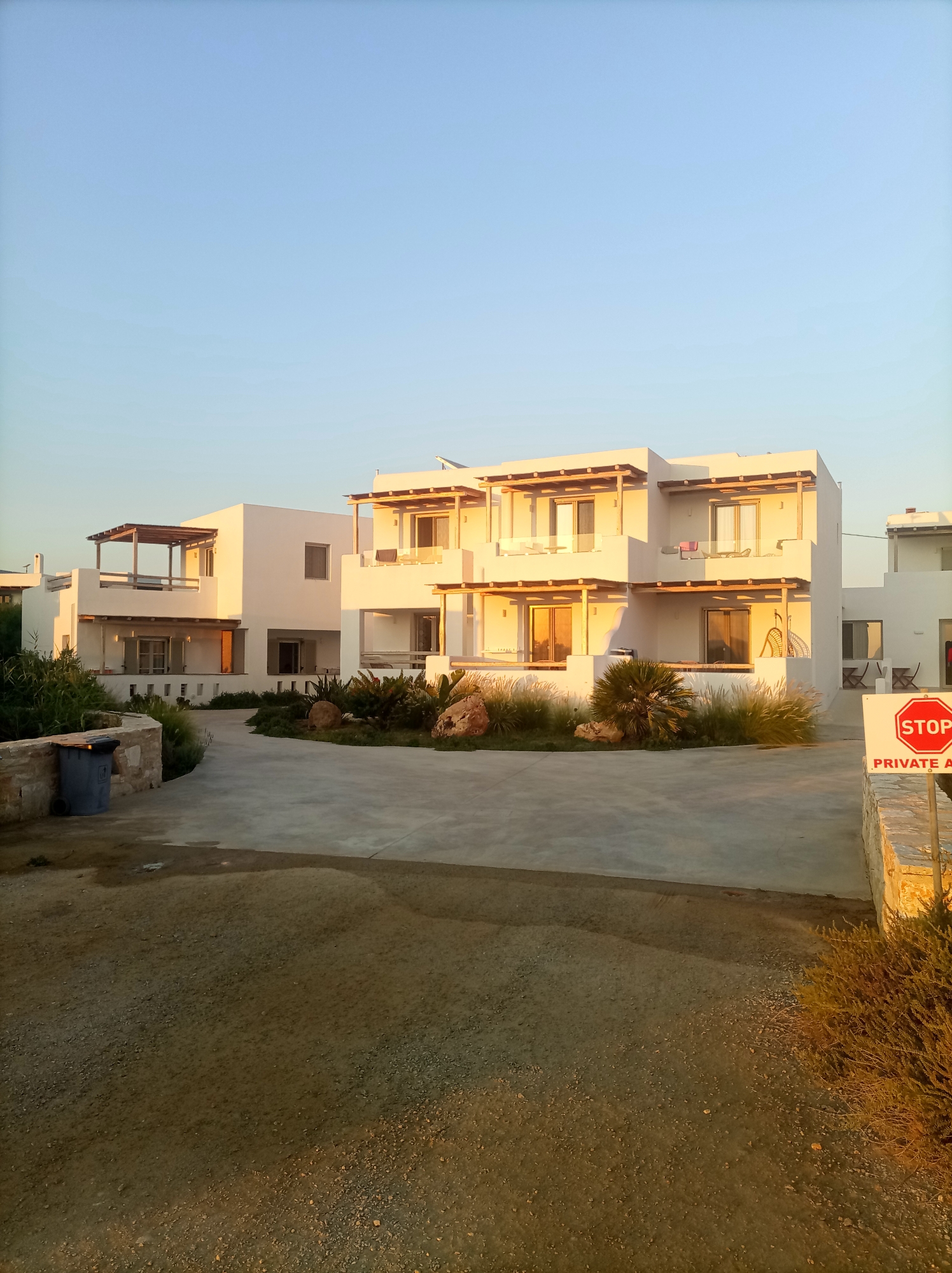In the city of Naxos, Greece, the image captures a modern architectural complex bathed in the golden light of a sunset. The buildings, painted white with exposed wooden beams on their balconies, present a clean and minimalist aesthetic. They are multi-story structures with multiple balconies and large glass doors, suggesting they are likely apartments or a small hotel.
In the foreground, a paved driveway curves around to the buildings. To the left, a dark blue garbage bin sits beside a low stone wall. Further into the scene, a variety of drought-tolerant plants and grasses are arranged around the buildings, some with large rocks interspersed among them. A sign with a red stop symbol and the words "PRIVATE A" is prominently displayed on the right, indicating restricted access. The sky above is a clear, pale blue, transitioning to a warmer hue near the horizon. The overall impression is one of a serene and well-maintained property in a Mediterranean setting.