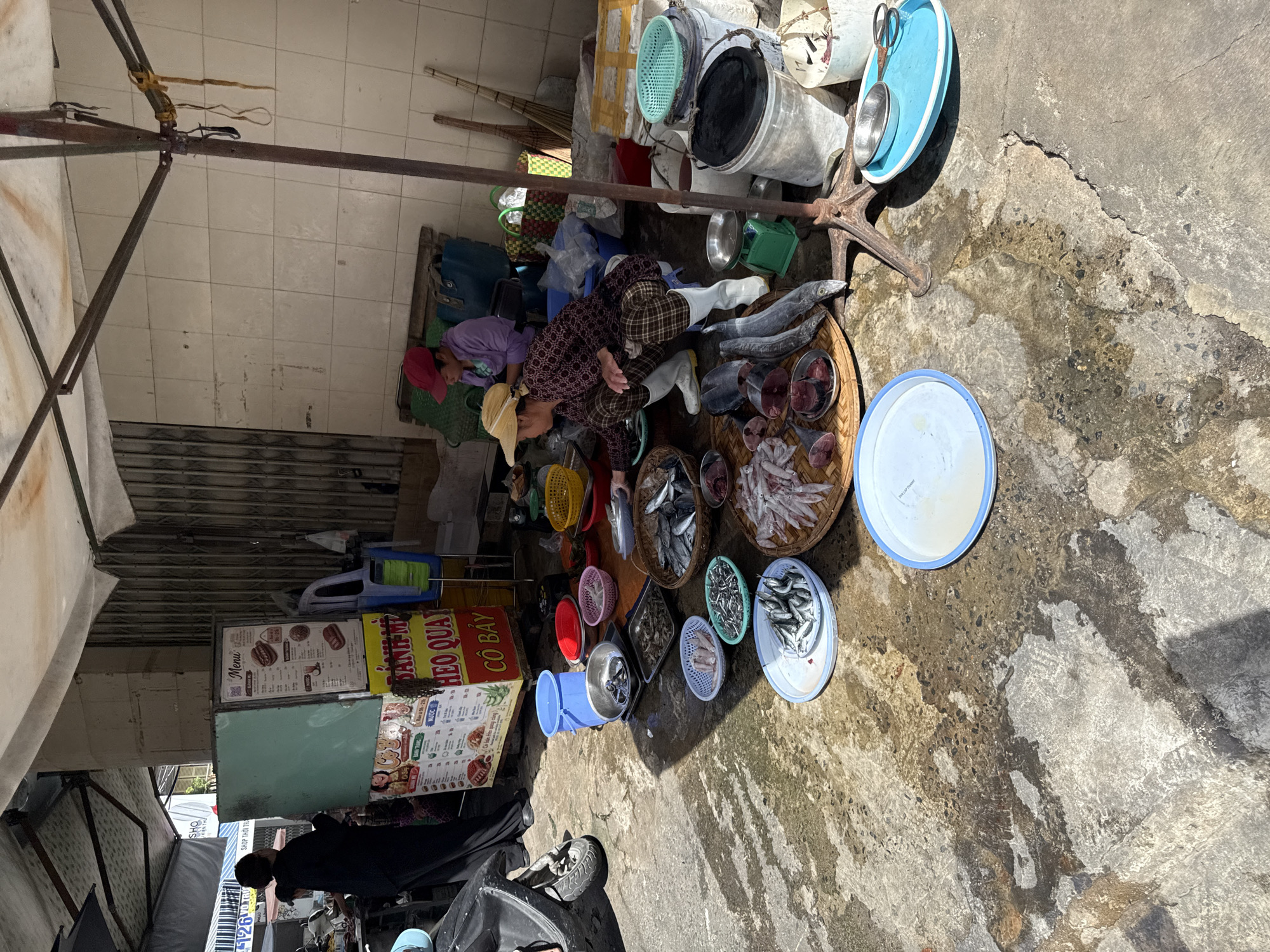 The image captures a street market scene in Nha Trang, Vietnam, likely on a sunny day, judging by the strong shadows. The foreground is dominated by a wet, concrete ground surface with patches of light and dark, indicating water or oil spills. Scattered across the ground are various food items, primarily fish. There are several woven baskets and plastic trays containing different types of fish, some whole and some cut into pieces. Some of the fish appear to be prepared for sale, with scales removed or heads severed. There are also several metal bowls and plates, as well as a small green weighing scale.
In the mid-ground, two women are visible. One woman, wearing a straw hat and a patterned shirt, is crouching down and appears to be arranging or preparing fish on a woven tray. She is wearing plaid pants and white boots. To her left, another woman in a purple shirt and a red cap is seated, also engaged in activity related to the market. Behind them, a turquoise refrigerator is visible, with a menu board displaying food items, suggesting it is part of a food stall. A motorbike is parked on the left side of the frame.
The background features a stall with a white canopy, supported by metal poles. A corrugated metal wall forms the backdrop to the stalls. Various items, including plastic buckets, bags, and what appear to be brooms, are present. The overall impression is a bustling, informal market environment where fresh produce, specifically fish, is being sold.