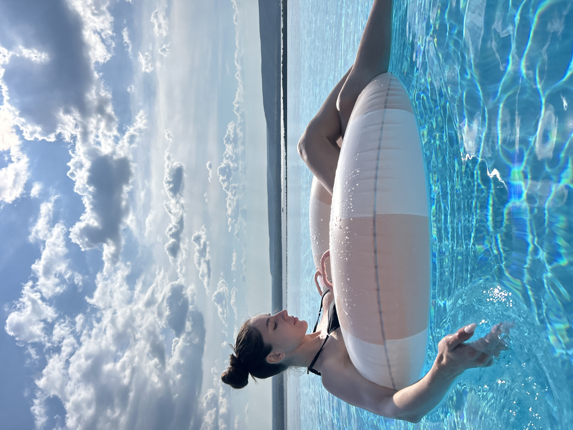 A woman with dark hair styled in a bun is floating in a clear blue infinity pool on an inflatable ring. She is wearing a black bikini. Her body is partially submerged in the water, and her legs are extended. Her left hand rests on the inflatable ring, with her fingers trailing in the water. The background shows a calm body of water, likely a reservoir, with rolling hills in the distance. The sky is filled with dramatic clouds, with sunlight breaking through in some areas. The scene appears to be a leisurely moment of relaxation in an outdoor pool setting. The location is identified as Novokuybyshevsk, Russia.