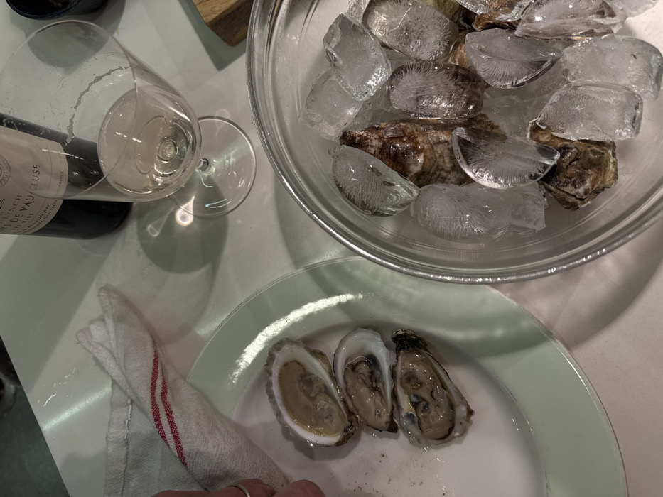 The image displays an indoor scene in New York, United States of America, focused on food and beverage preparation. On the left side, a partially visible hand, wearing a silver-toned ring on the ring finger, is positioned near a white plate with a light green rim. Three shucked oysters rest on this plate, showing their internal contents. A white cloth napkin with two thin red stripes is folded beside the plate. To the right, a clear glass bowl is filled with unshucked oysters covered by multiple ice cubes. In the upper right section, a dark glass wine bottle, bearing a label that partially reads "CH. LYNCH" and "DE VAUCLUSE", stands upright next to a stemmed wine glass containing a pale yellow liquid, presumably white wine. All items are arranged on a smooth, light-colored surface, which reflects light and casts shadows from the glass and bottle.