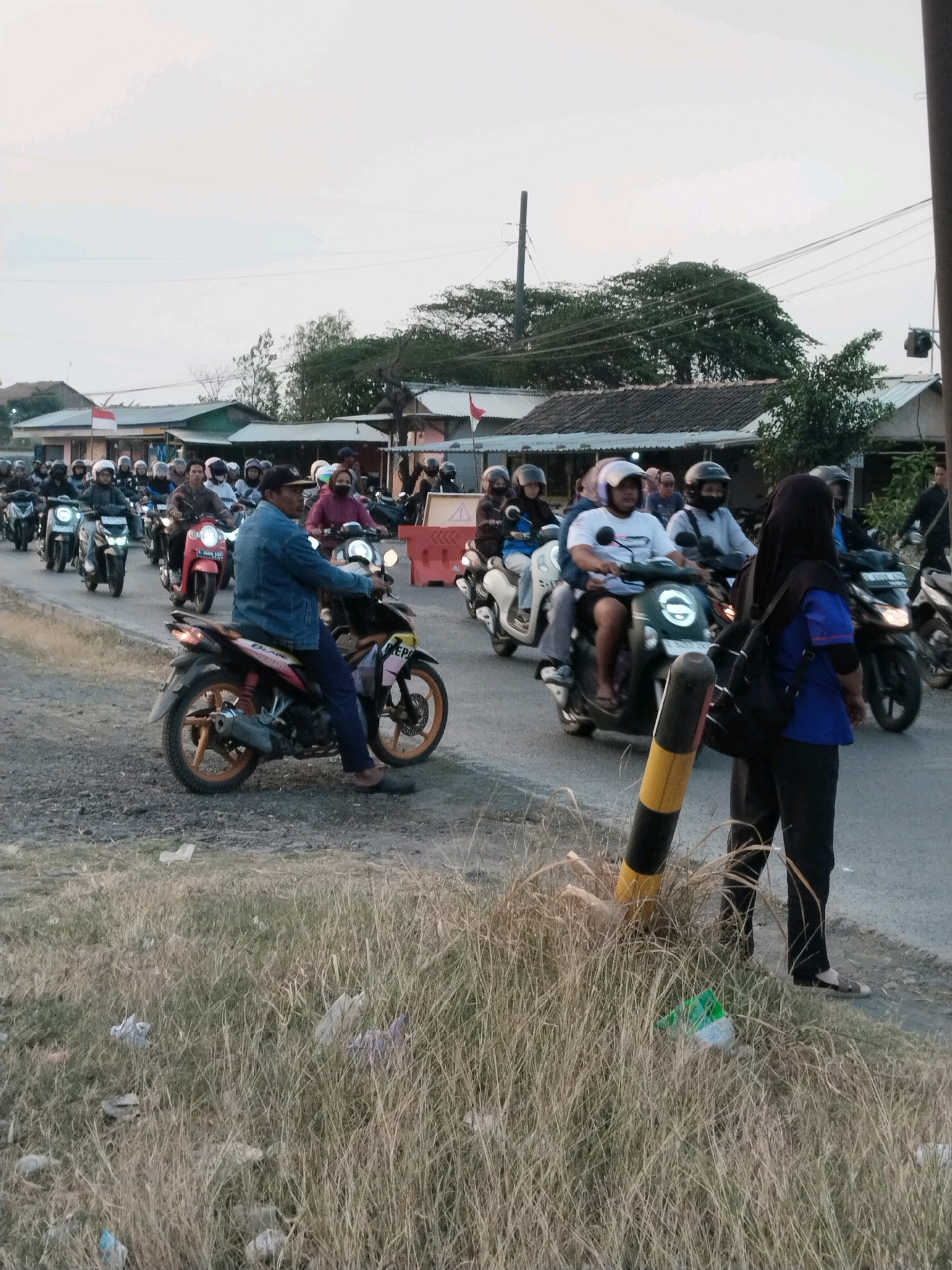 The image depicts a street scene in Songgom, Indonesia, with a line of motorcycles forming a procession. In the foreground, a man wearing a denim jacket and blue pants rides a motorcycle with orange rims, looking towards the left. He is in the right lane, and the motorcycle has the license plate "E 4609 ABM". To his right, a woman wearing a black headscarf and a blue shirt is visible from the back, carrying a backpack and facing away from the camera, standing near a traffic bollard. Behind them, a multitude of motorcycles, many with riders wearing helmets, are traveling in the same direction. The motorcycles vary in color and model, with some appearing to be scooters and others more traditional motorcycle types. The environment includes buildings with corrugated metal roofs lining the street, some adorned with Indonesian flags. Trees and vegetation are visible in the background, suggesting a semi-urban or rural setting. The ground on the left side of the street is covered in dry grass and some scattered litter. The sky is overcast, indicating either dawn or dusk.