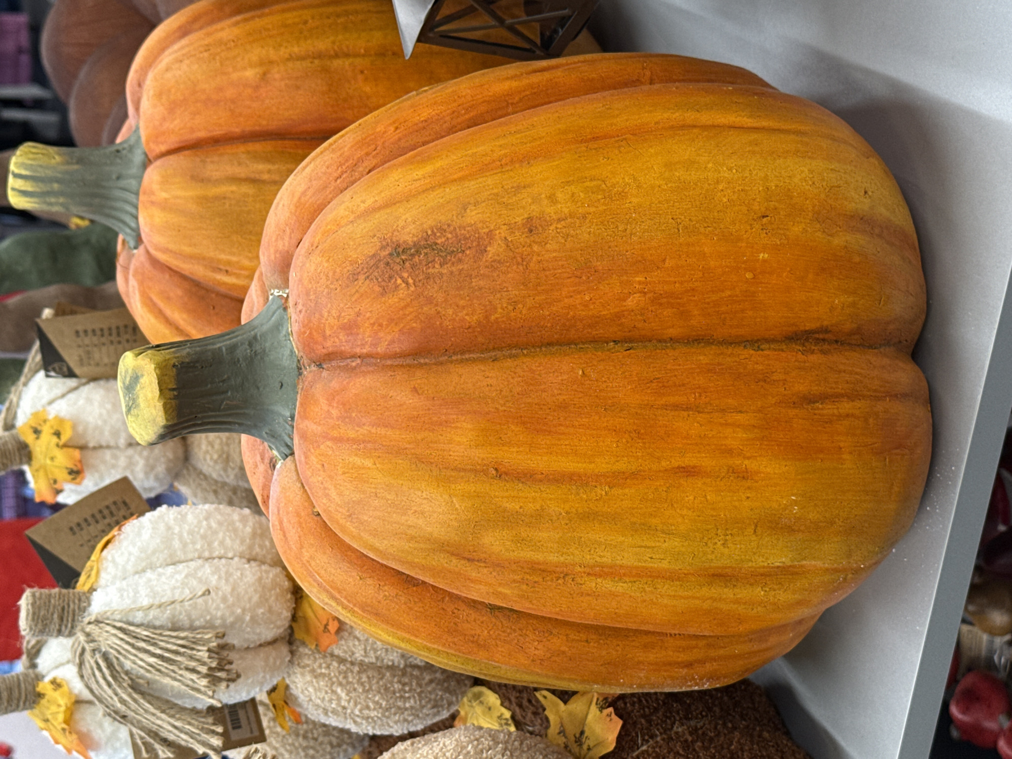 The image shows a close-up of several artificial pumpkins displayed on a shelf. The pumpkins are of various sizes and colors, with the dominant pumpkins being large and orange with textured surfaces and green stems. In the background, there are smaller, white, fuzzy pumpkins adorned with jute twine and artificial leaves. The overall scene suggests a decorative display, likely for autumn or Halloween. The image is taken from a slightly elevated angle, focusing on the pumpkins. There is no indication of the location being in Bad Lauterberg im Harz, Germany.