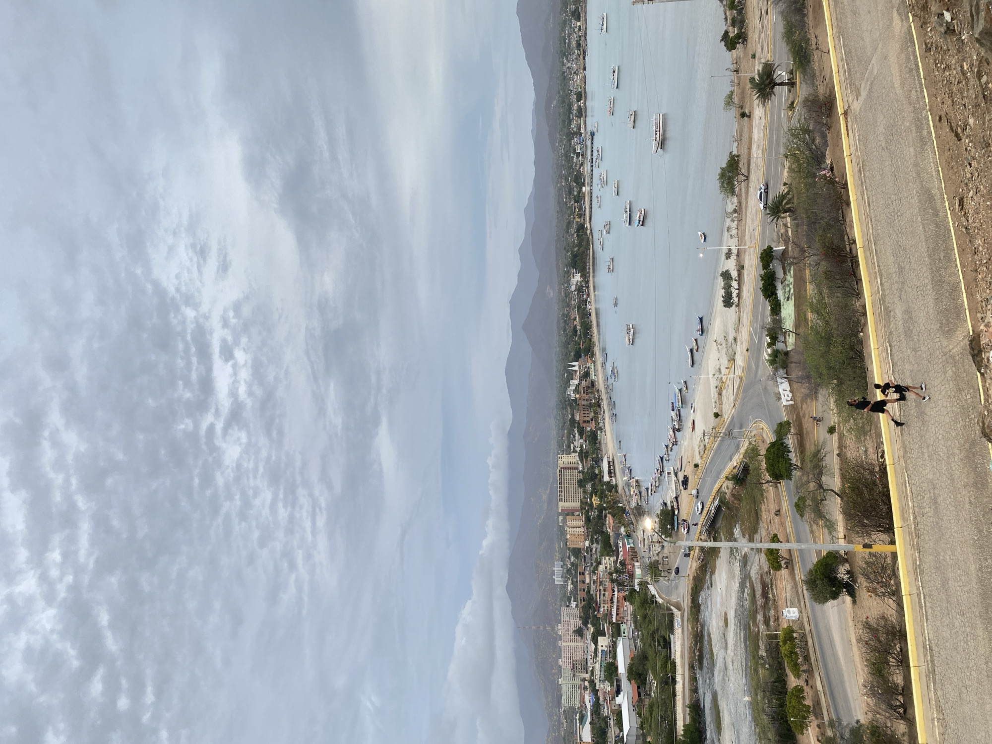 The image provides an elevated, wide-angle view of the coastal city of Juan Griego, Venezuela, under a heavily overcast sky. The sky is uniformly covered with light grey, textured stratocumulus clouds.
In the distant background, a range of undulating, green-grey mountains extends across the horizon, partially obscured by atmospheric haze. Below these mountains, the urban landscape of Juan Griego unfolds, featuring a dense arrangement of buildings, including multi-story residential and commercial structures, along with smaller houses. A prominent white church with twin steeples is visible on the left side of the town, near the bay.
The central feature of the midground is a calm body of water, likely a bay or inlet, populated by numerous boats of varying sizes, predominantly smaller recreational or fishing vessels, many with white hulls. A paved road curves along the shoreline of the bay, where several vehicles are present. Adjacent to this coastal road, a sandy strip of beach is visible, transitioning into an area of sparse, dry vegetation and what appears to be a marshy or tidal flat section. A bridge spans a narrow waterway or dry creek bed that connects to the bay.
In the immediate foreground, a paved road ascends gently towards the left. Two individuals are depicted jogging downhill on this road, both wearing dark athletic attire. One individual is positioned slightly ahead and to the left of the other. A yellow curb delineates the edge of this foreground road. The landscape surrounding the roads is characterized by dry, scrubby vegetation, small trees, and exposed earth. A tall streetlamp with a yellow pole stands on the lower coastal road, illuminating the area. Palm trees are also visible at various points along the coastal strip.
