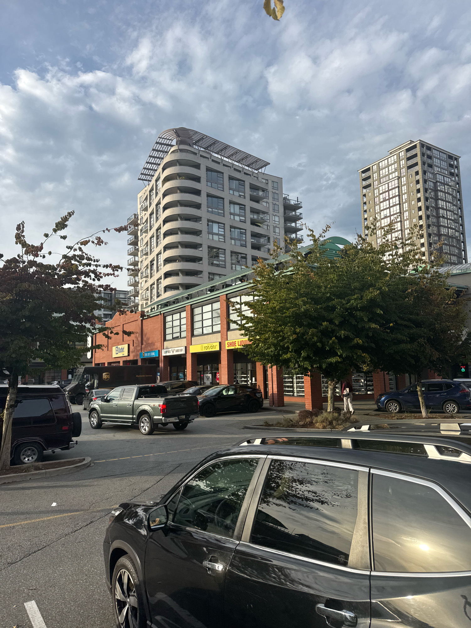 The image depicts a street scene in New Westminster, Canada, with a focus on urban architecture and vehicles. In the foreground, a dark-colored SUV is parked, its side reflecting the sky and trees. To its left, a dark-colored van is also parked. Moving towards the mid-ground, a silver pickup truck is driving away, and several other cars are visible on the street and in parking spots.
Dominating the background are several modern buildings. A curved, multi-story residential building with numerous balconies is prominent, topped with a sloped, metallic structure. To its right stands a taller, rectangular high-rise building with many windows. Below these residential towers, a strip of retail businesses with large windows faces the street. Signs visible include "Outlet," "The UPS Store," "ron rons," and "SHOE LIQUOR." Trees line the sidewalk in front of the businesses, some with fall foliage. A person in white pants and a dark top is walking on the sidewalk. The sky is partly cloudy, with the sun breaking through in some areas, casting reflections on the vehicles.