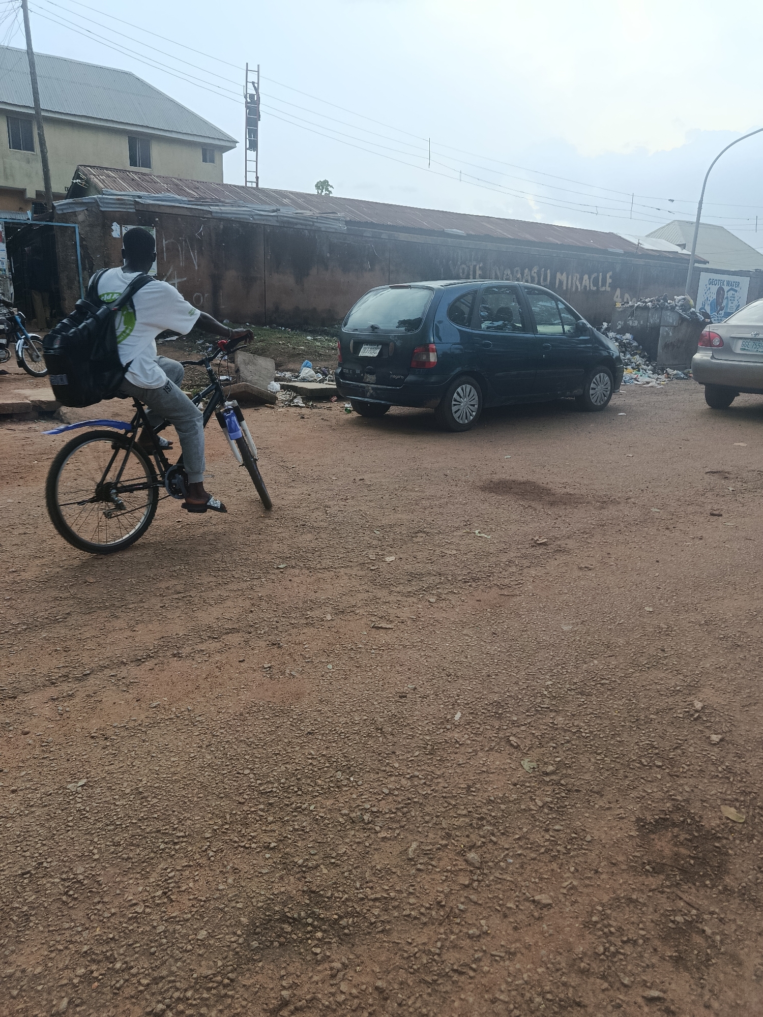A young Black male wearing a white t-shirt, grey pants, and sandals rides a bicycle with a large black backpack. He is on a dirt road in what appears to be a dusty, urban environment. In the background, there is a dark blue minivan, a silver car, and a long, corrugated metal building with graffiti. A trash-filled area is visible next to the building. The sky is overcast. This image was taken in Rafin Par, Nigeria.