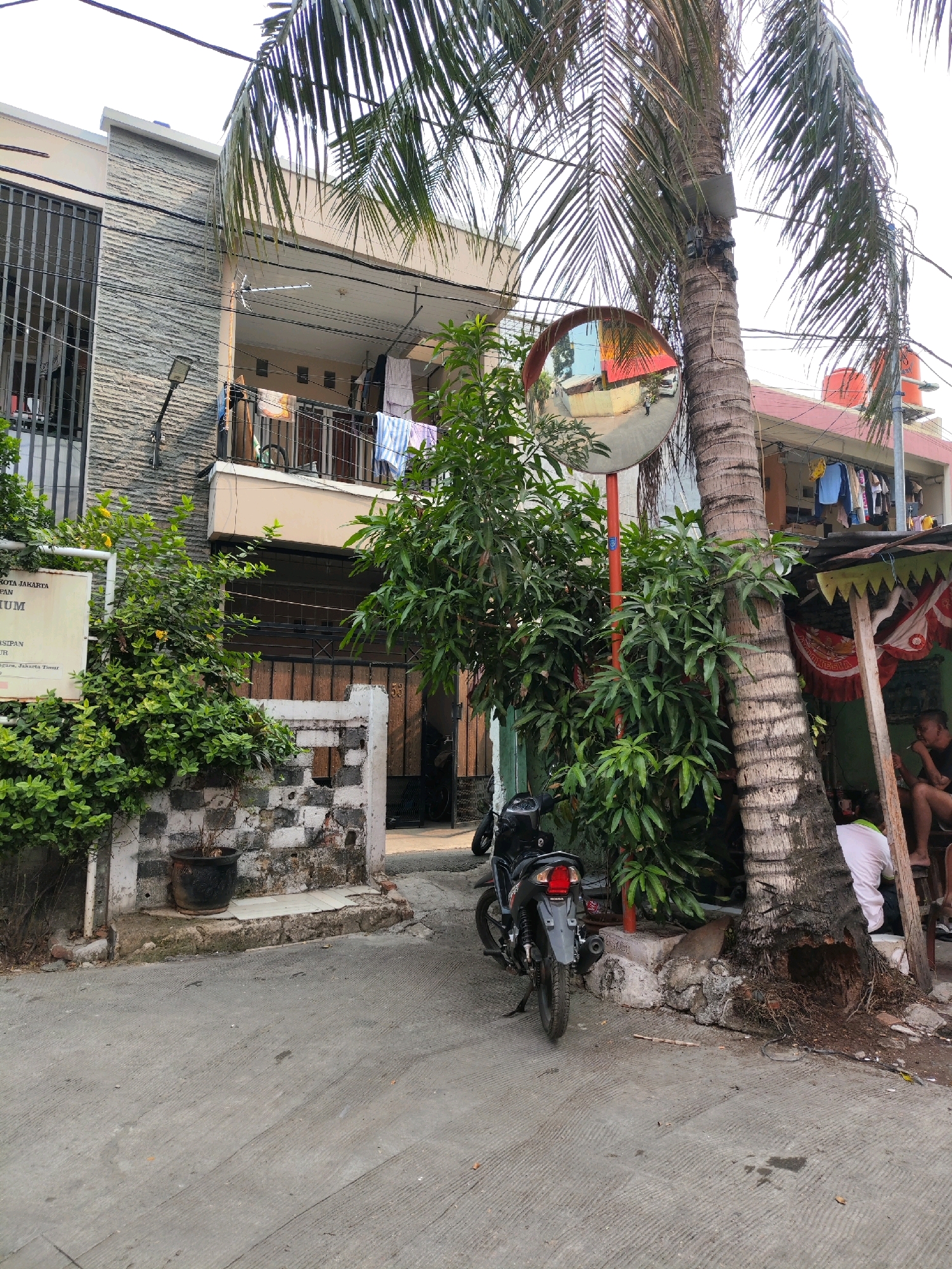 The image depicts a street scene in the city of East Jakarta, Indonesia. In the foreground, a gray asphalt road leads into the frame. To the left, a multi-story building with a gray stone facade and dark metal bars on the windows is visible. A white sign with text in Indonesian is mounted on the building. Lush green bushes and plants are growing around the base of the building.
A black motorcycle is parked on the left side of the road. Further into the image, a large palm tree with a textured trunk stands prominently. Attached to the palm tree is a convex traffic mirror mounted on a red pole, reflecting the street. Behind the palm tree, another building with a red roof and several windows is partially visible, with laundry hanging on a balcony.
To the right of the palm tree, a small, makeshift structure with a green and yellow awning is set up. Two individuals are visible in this area. One person is wearing a white shirt and is bent over, facing away from the camera. Another person, partially obscured, is seated and smoking a cigarette. The ground is a mixture of concrete and dirt, with exposed tree roots from the palm tree. The overall environment suggests a residential or commercial area in a tropical urban setting.