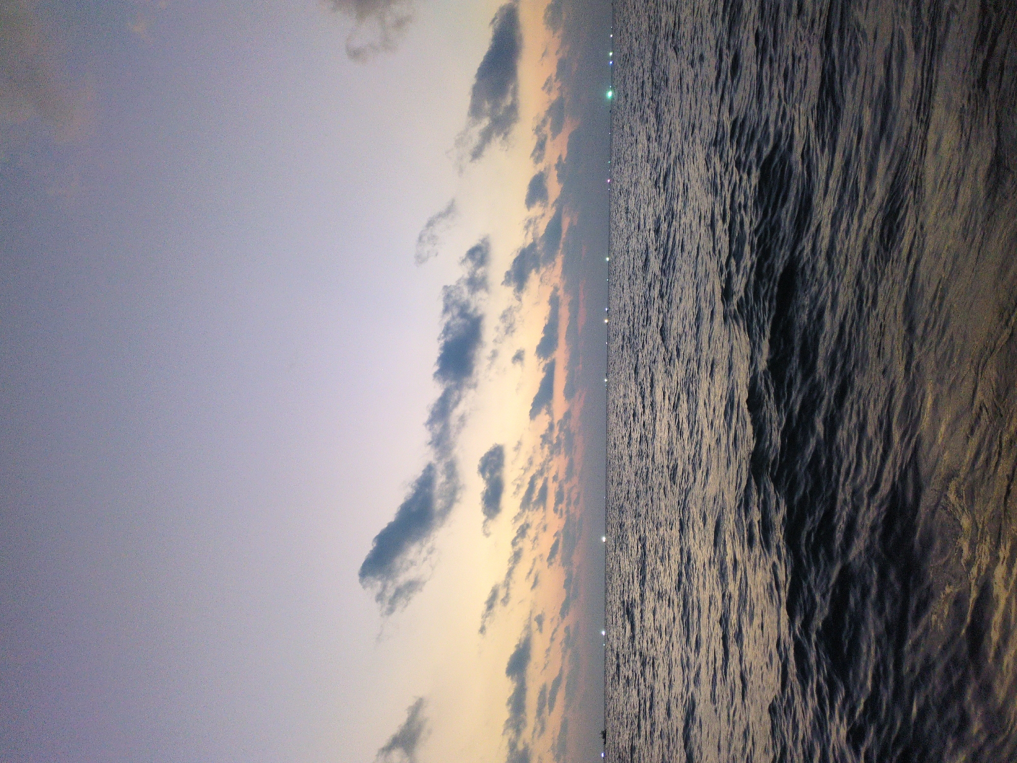The image shows a landscape view of the ocean at sunset in Phú Quốc, Vietnam. The sky is divided into two halves; the left is a grayish-purple sky and the right is filled with orange, pink, and gray clouds. The sea occupies a substantial portion of the image, exhibiting a rippled texture. The presence of lights along the horizon suggests a coastal road or city at night.