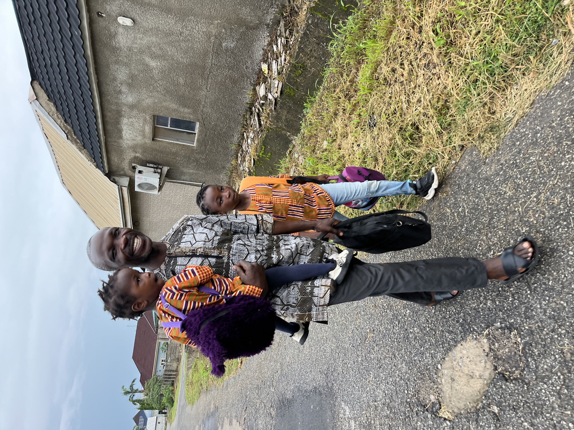 An upright-oriented, full-length shot shows an adult male with two young girls walking outdoors on a paved road under an overcast sky. The man, with short grey hair and a beard, smiles broadly at the camera. He wears a patterned black and white traditional African shirt, dark trousers, and black open-toed sandals. He carries a black shoulder bag on his left side and holds a young girl in his left arm.
The younger girl, held by the man, wears an orange and brown patterned top, dark leggings, and has a fuzzy purple bag slung over her shoulder. She looks up towards the man. To their right, an older girl walks independently, wearing a similar orange and brown patterned top, light blue jeans, and black and white sneakers. She carries a purple backpack and looks directly at the camera.
The background features a grey concrete building with a dark tiled roof on the left, possibly a house, featuring a small window and an external air conditioning unit. The right side of the frame shows a strip of overgrown grass and weeds separating the paved road from what appears to be a concrete wall or gutter. The paved road has a small pothole near the man's feet. The overall impression is a casual outdoor moment, possibly a family outing in Abuja, Nigeria, given the architectural style and attire. There is no visible text.