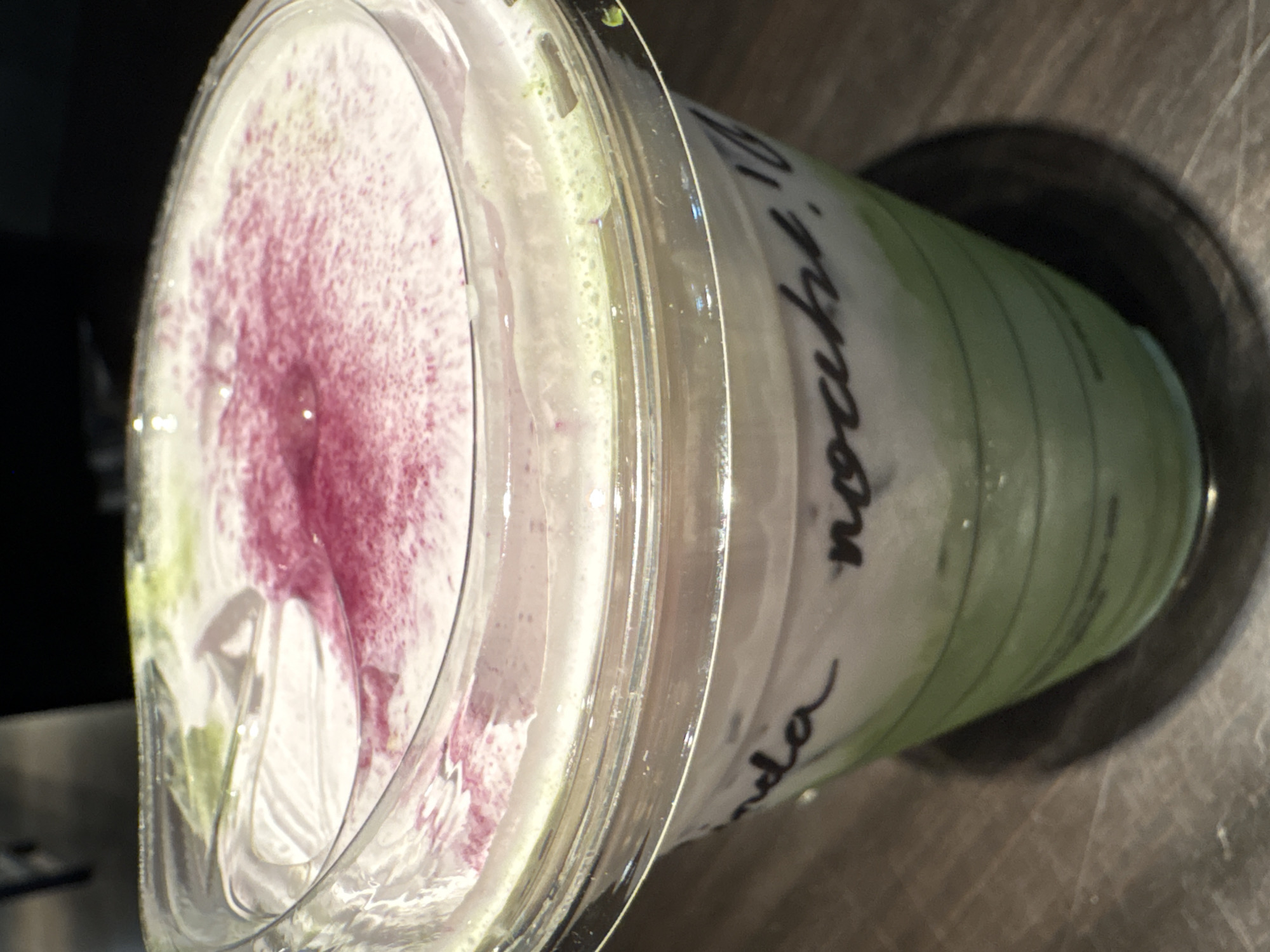 A slightly tilted top-down close-up photograph captures a cold beverage in a clear plastic cup with a lid. The cup rests on a dark, possibly metal, circular surface. The beverage inside appears layered; green at the base, transitioning to a white, creamy layer, and topped with a pink, speckled foam. There is writing in black marker on the side of the cup, partially obscured, but "nocuhi" is clearly visible. The setting appears to be a coffee shop or cafe, possibly in Mexico City, Mexico, based on the drink and potential location. The image is well-lit, with some reflections on the plastic cup.