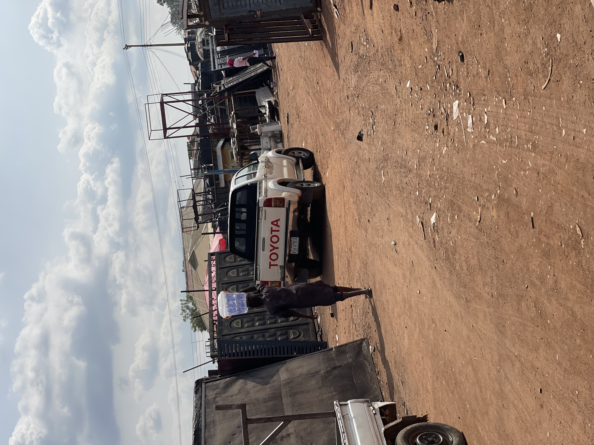 A medium, outdoor shot depicts a dirt-covered area next to what appears to be a metalworking or fabrication yard in Abuja, Nigeria. A white Toyota pickup truck is parked, with its rear facing the camera. A dark-skinned person is walking away from the truck, carrying a white container on their head. The yard to the left is filled with metal structures, sheets, and fabricated gates. Overhead power lines are visible against a partly cloudy sky. The ground is brown and sparsely littered.