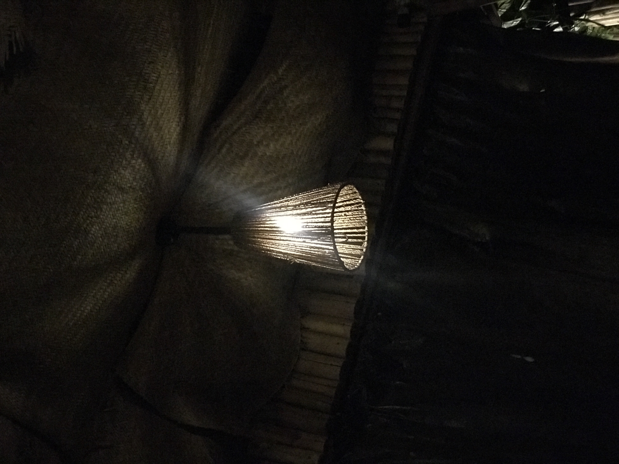 A dimly lit interior scene features a woven lampshade emitting light. The light is pointed towards a woven wall surface. To the right, a darker area suggests a corner or edge of a structure, possibly made of wood. It is impossible to determine the location of Abuja, Nigeria, or the presence of people or specific actions from the visual data.