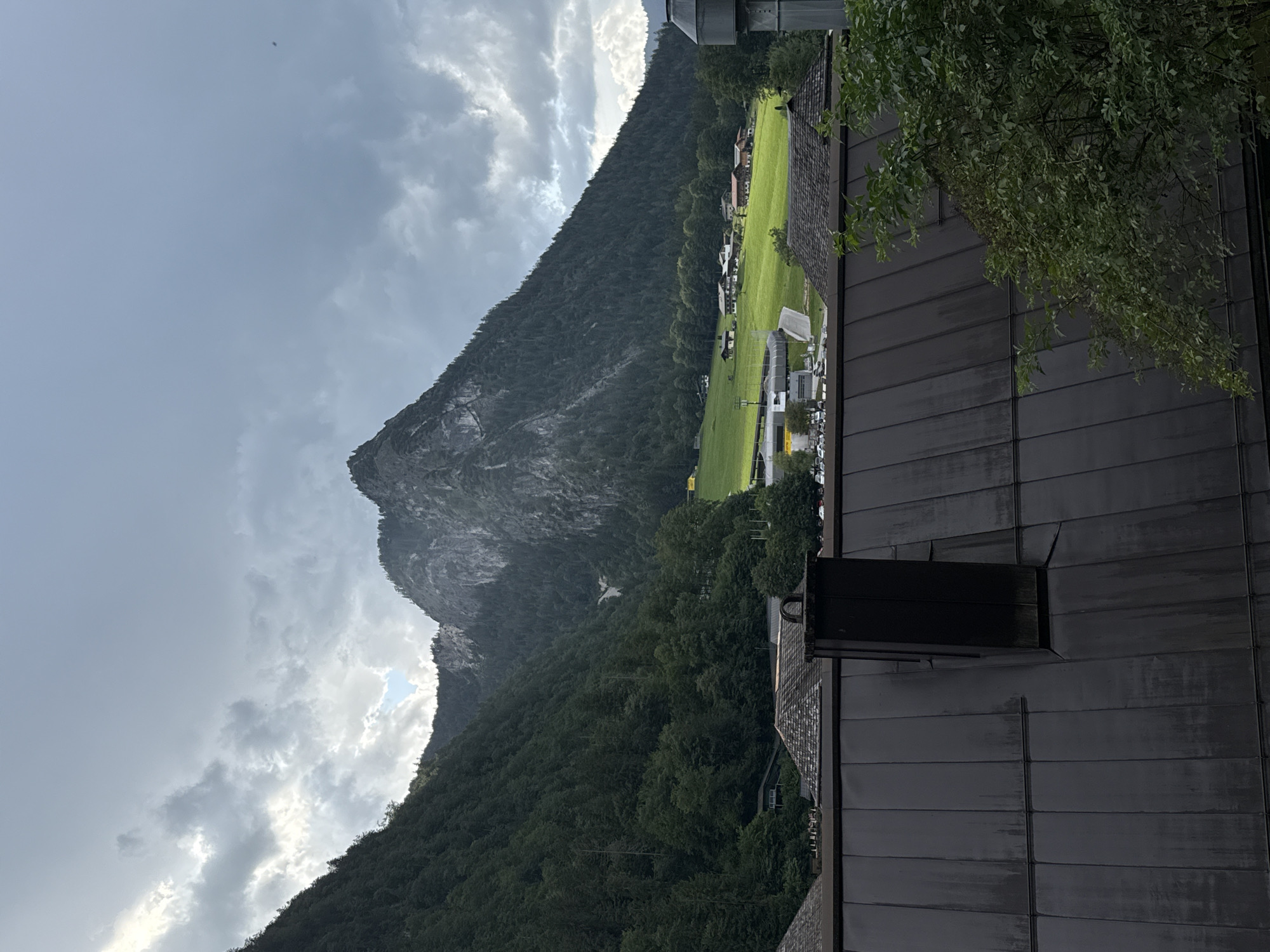 The image captures a view of Schönau am Königssee, Germany, dominated by a towering, rugged mountain face on the left, densely covered in dark green conifer trees. The mountain slopes descend towards a lush green valley dotted with several traditional Bavarian-style buildings, some with red roofs. A bright yellow section, possibly a field or sports facility, is visible in the middle ground. The sky is overcast with dramatic, dark gray clouds, interspersed with patches of lighter sky, suggesting an approaching storm or recent rain. In the foreground, a dark, metallic roof with a visible chimney is prominent. A section of a vibrant green tree with dense foliage is visible on the right side of the frame. The overall scene evokes a sense of mountainous terrain and rural Bavarian architecture under a moody sky.