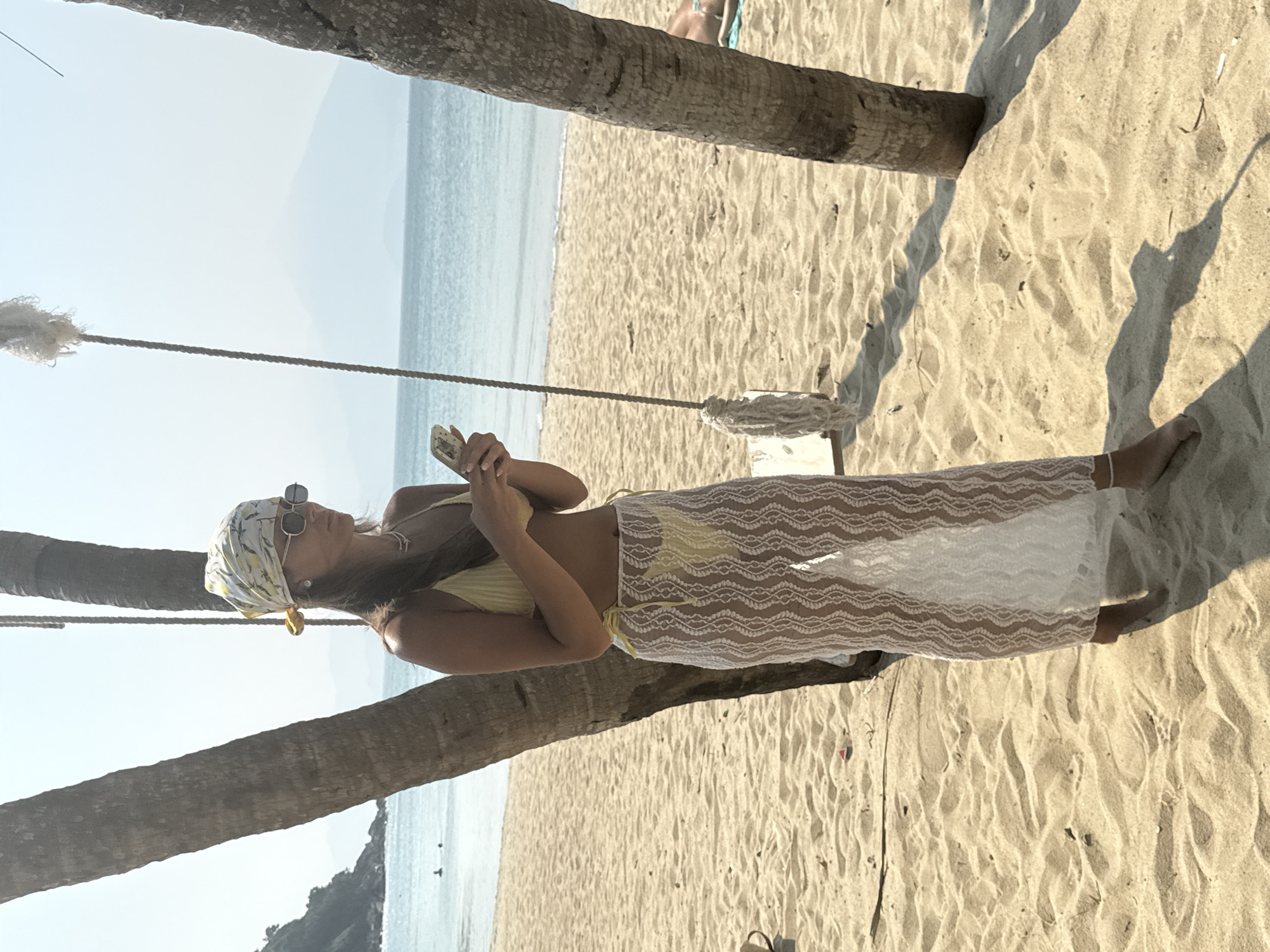 A woman stands on a beach at Đà Nẵng, Vietnam. She is wearing a yellow bikini, a white lace sarong, sunglasses, and a headscarf. She is holding a phone in her hands. A swing hangs from a palm tree. The ocean is visible in the background.