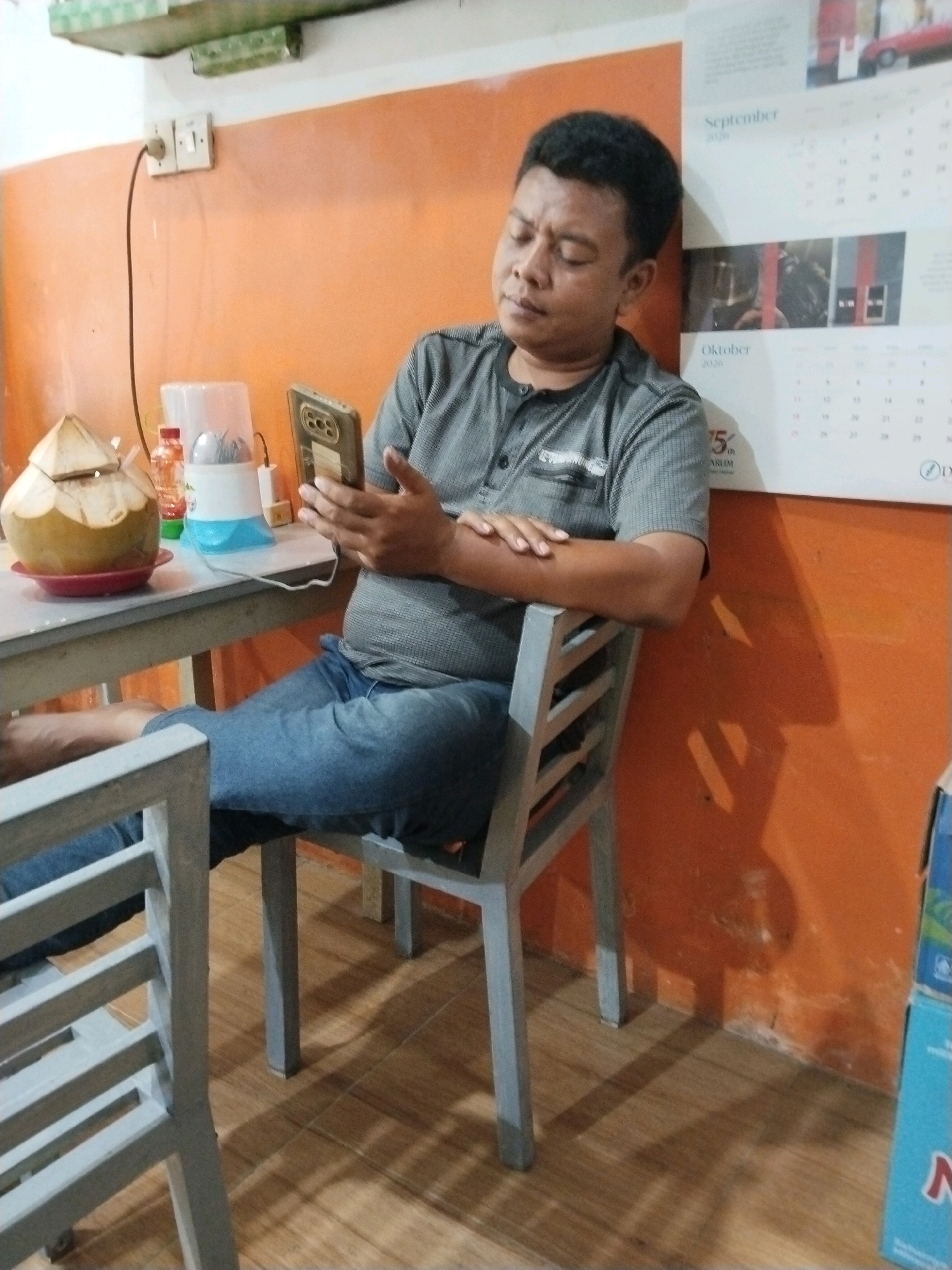 A man sits at a table in an orange-walled room, likely a restaurant or warung in Prigi, Indonesia. He is wearing a grey, short-sleeved polo shirt and blue jeans. The man is looking down at his smartphone, holding it in his right hand. His left arm is resting on the back of the metal chair he is sitting on. On the table in front of him is a halved coconut with a straw inserted. To the right of the coconut are a clear plastic bottle with a red lid and a white appliance with a blue base. Behind the man, a calendar with the months of September and October 2020 is hanging on the wall, along with some framed pictures. The floor is tiled with light brown wood-patterned tiles. The overall lighting is dim, with shadows cast by the objects and the man.
