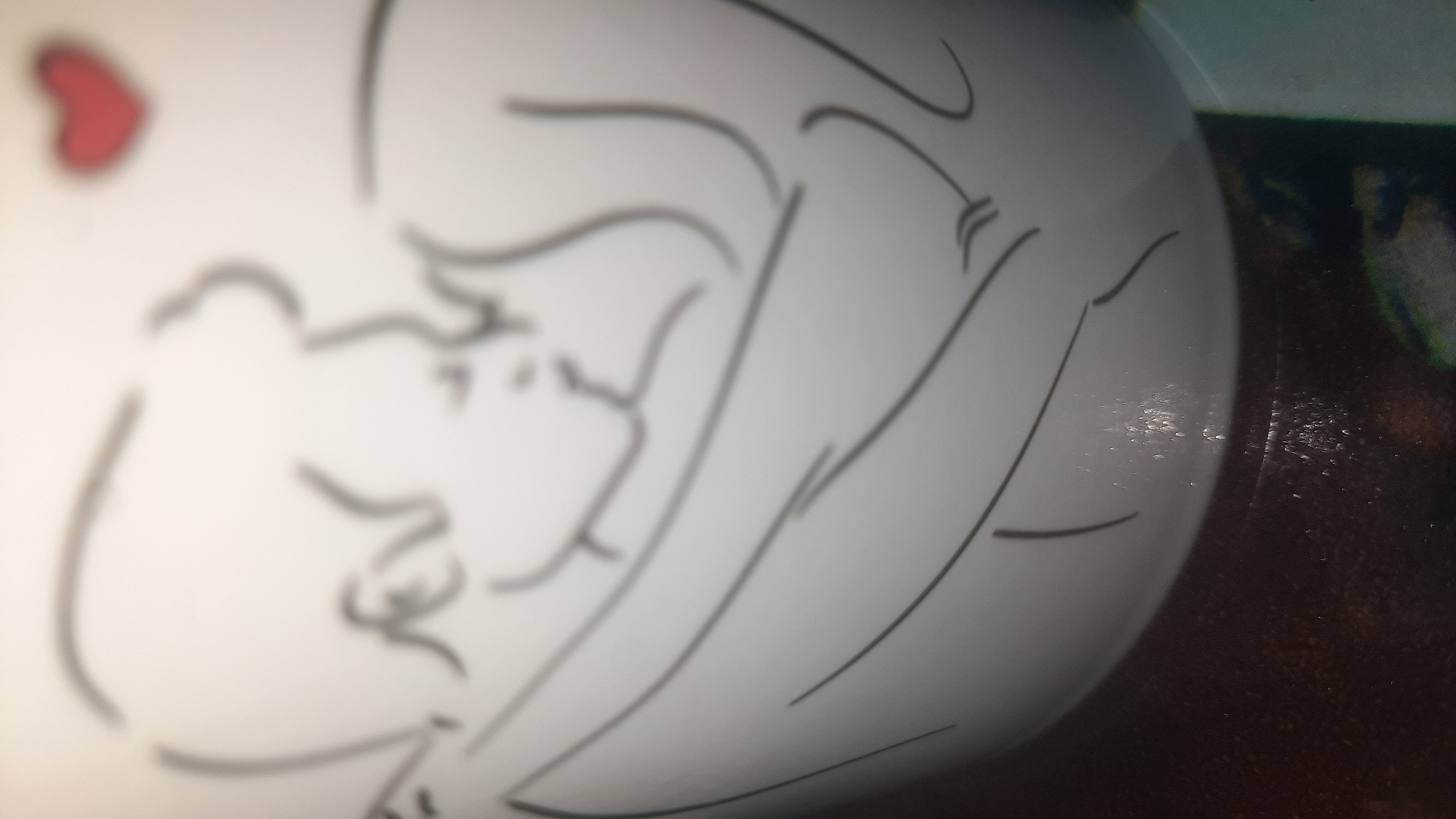 The image is a close-up, slightly blurred photograph of a white ceramic object, possibly a mug or a vase, with a black line drawing on its surface. The drawing depicts two figures in an embrace, with one figure appearing to lean their head towards the other. Above the figures, a small, red heart is drawn. The ceramic object is curved, suggesting it is a rounded surface. The lower portion of the image shows a dark, textured surface, likely a table or countertop, with some reflective water droplets or condensation on the ceramic object. The background is out of focus, but appears to be a neutral color. There is no visible context indicating a specific location in the city of Шклоў, Belarus, or any other identifiable urban setting.