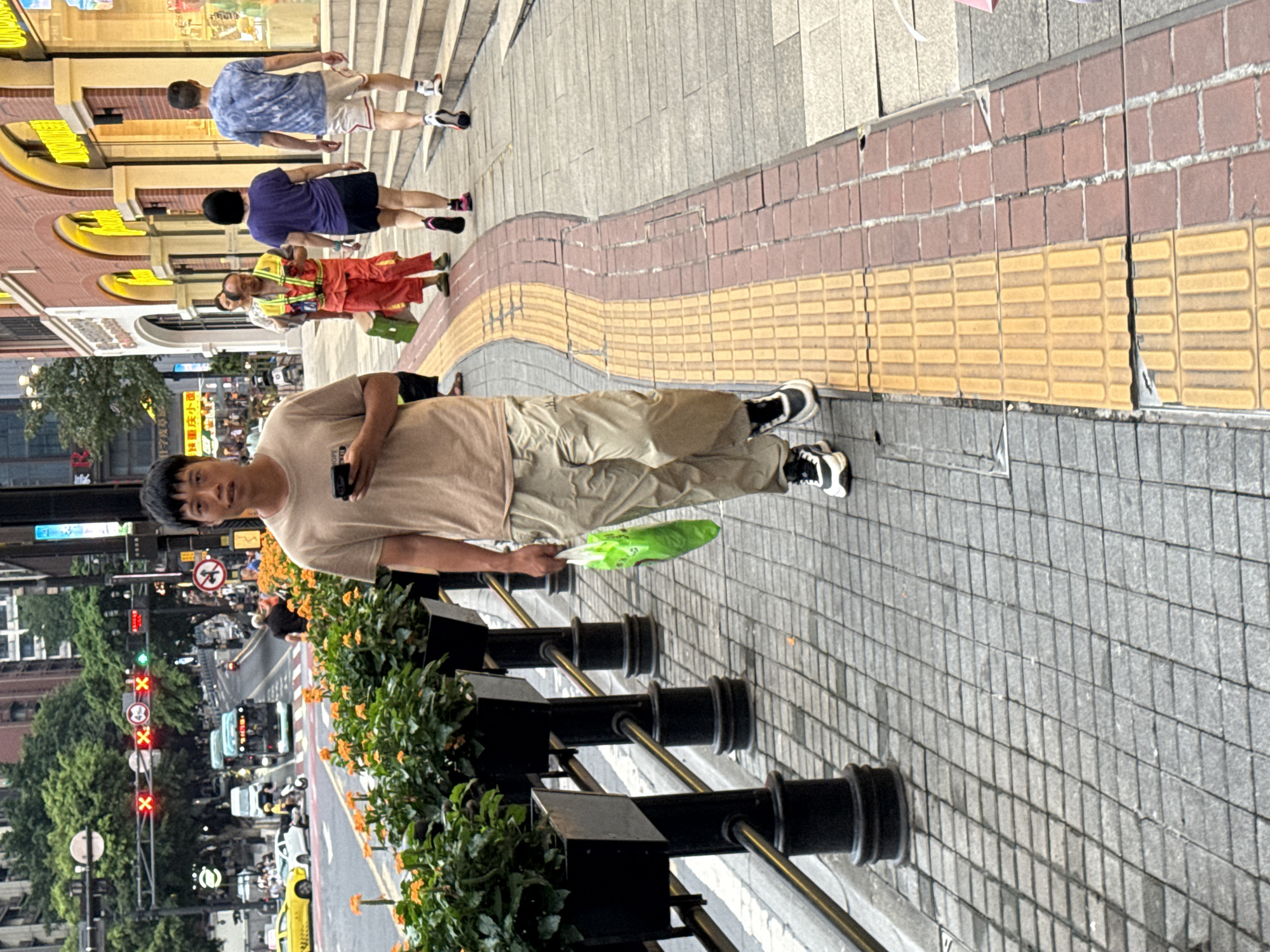 A man walks on a sidewalk in Yuzhong District, China, holding a mobile phone and a green plastic bag. He is wearing a tan t-shirt and khaki pants with black and white athletic shoes. The sidewalk has a textured yellow and red brick pattern. In the background, there are buildings with arched windows, a street with traffic including a yellow taxi and a bus, and planters with orange flowers. Other pedestrians are visible further down the sidewalk, including a person in a bright orange vest and red pants. The scene appears to be in a commercial or urban area.