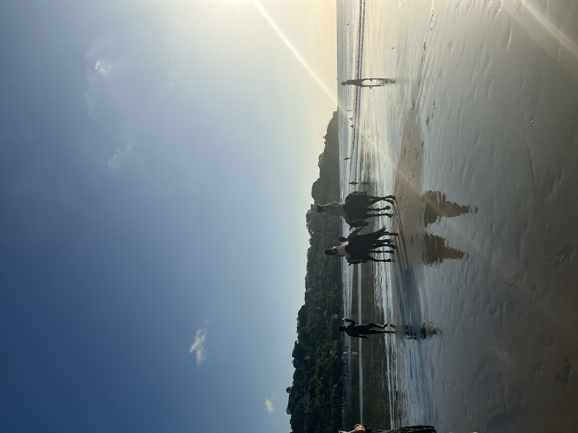 In Kuta Selatan, Indonesia, a beach scene unfolds under a clear blue sky with a faint hint of a distant cloud. The sun, positioned high and slightly to the right, casts a bright glare and a radiant sunbeam across the ocean and wet sand. The tide is low, leaving a broad expanse of reflective sand that mirrors the sky and the figures on it.
Several people are present. In the foreground, on the left, a person is partially visible, holding a phone. Slightly further into the scene, a lone figure walks away from the viewer, also holding a phone.
The main subjects are two horses with riders. The horse on the left is dark-colored, with a rider wearing a light-colored top. The horse on the right is also dark, with a rider dressed in a light-colored outfit. Both horses are walking towards the left of the frame. Their reflections are clearly visible on the wet sand.
In the mid-ground, several other individuals are seen walking or standing near the water's edge. Some appear to be swimming or wading. In the background, a lush, green hillside is dotted with buildings, suggesting a coastal settlement. The ocean waves are gentle, rolling onto the shore.