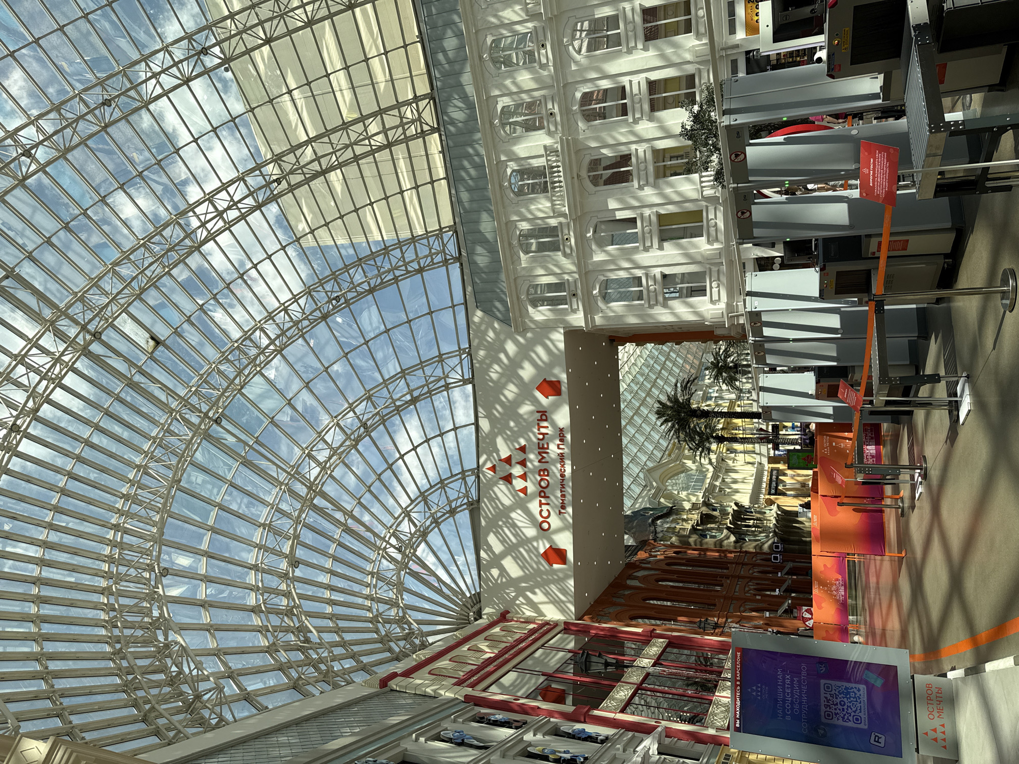 This is an interior shot of Ostrov Mecht, a theme park in Moscow, Russia. The view is looking upward, featuring a large arched glass and steel structure that forms the ceiling, allowing a view of the sky. Below the ceiling structure, there is a white building facade with multiple windows. A large sign is visible with the theme park's name, "ОСТРОВ МЕЧТЫ" in Russian, accompanied by arrows. The interior of the park is partially visible, including a row of palm trees and various architectural details. In the foreground are metal detectors, a QR code poster, and orange barricades.