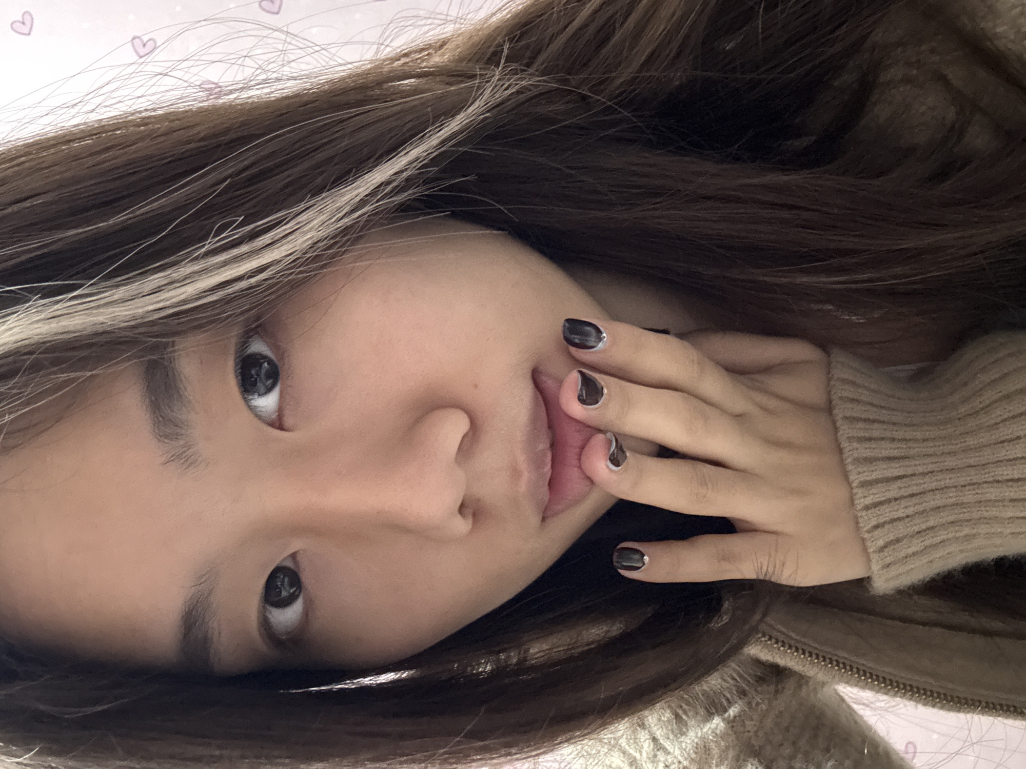 An Asian female with brown hair is positioned in the center. She wears a tan, zipped sweater. Her left hand is near her mouth, with dark nail polish featuring a silver stripe near the base. The background features pink hearts. It's difficult to determine the location as Shapingba District, China, due to the lack of identifiable landmarks.