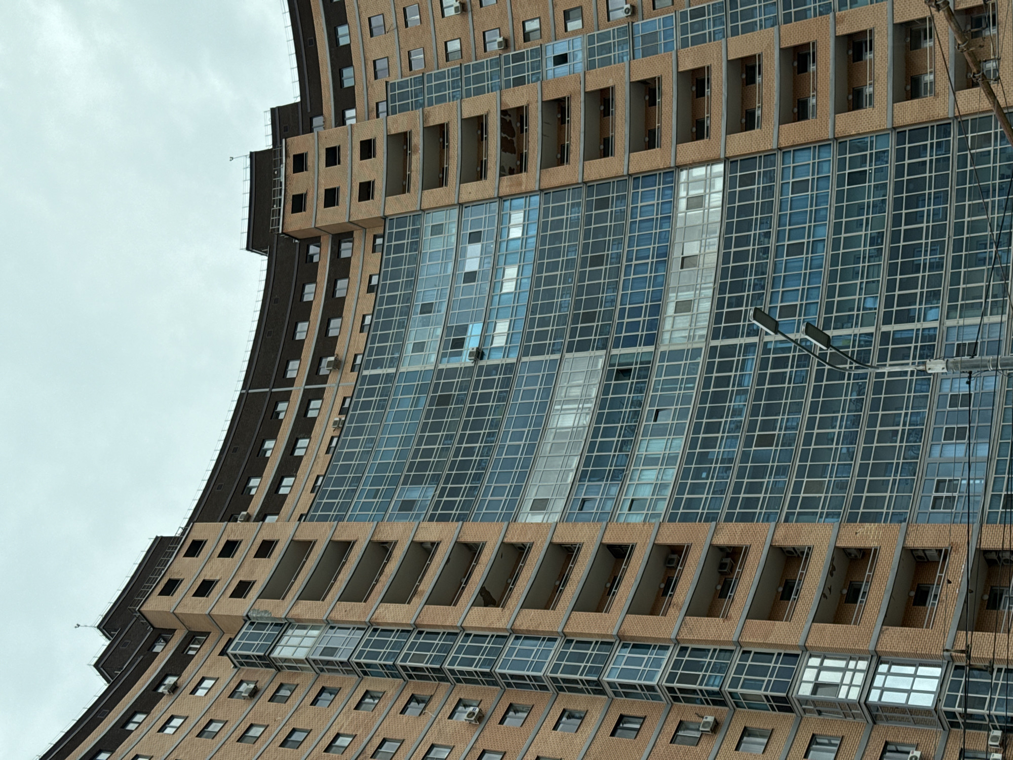 An exterior low-angle shot depicts a portion of an irregular-shaped, multi-story building with a modernist architectural style. The structure features a combination of brown brick and glass facades. The building's facade is segmented with alternating horizontal bands of recessed brick sections and expansive glass window panels. Some of the recessed sections house what appear to be exposed or open balconies. The windows are reflective, showing glimpses of the sky. The building curves in a distinctive manner, adding to its modern look.
The sky is overcast and grey. A few utility wires and streetlights are visible.
This building is located in Kazan, Russia.