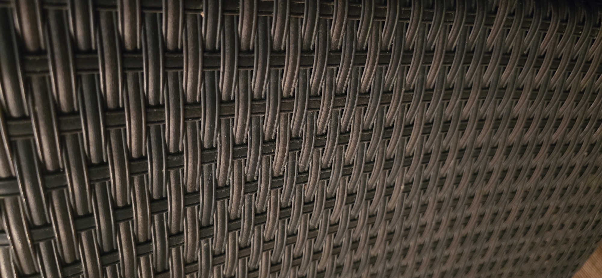 The image is a close-up, eye-level, vertical shot of a dark brown woven wicker or rattan surface. The weave consists of horizontal strands passing over and under vertical strands, creating a textured, patterned effect. The lighting appears to be artificial and casts some subtle shadows within the weave, highlighting the three-dimensional texture. The surface is uniform and shows no signs of damage or wear. The background is also filled with the same woven material, suggesting it is part of a larger object or structure like furniture or an outdoor seating area. There are no people, animals, or any other discernible objects in the image. The location is not discernible from the image itself.