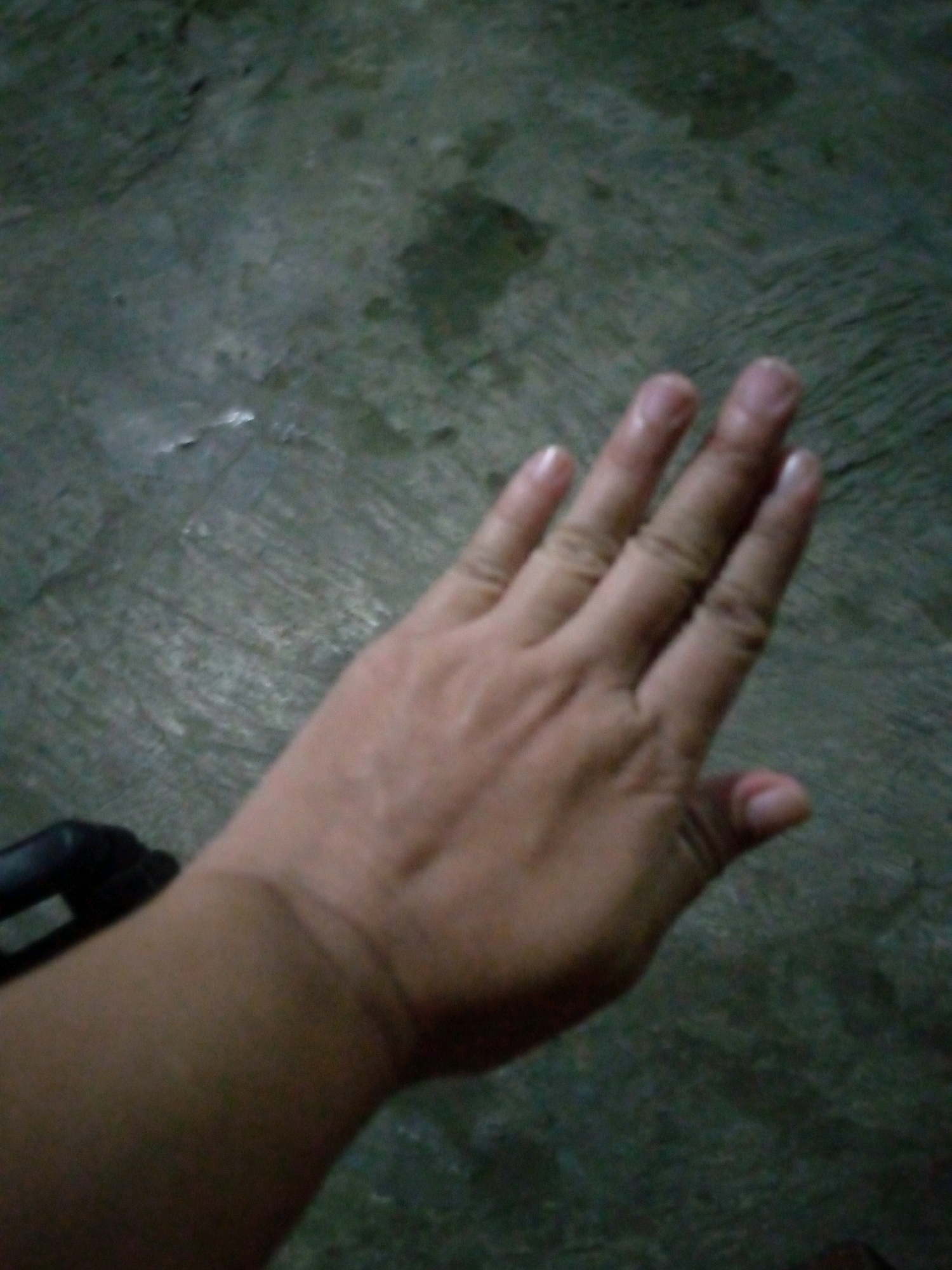 A person's hand is visible in the foreground, with the palm facing up and fingers slightly curled. The hand is positioned over a dark, textured surface that appears to be concrete or asphalt. A dark object, possibly part of a railing or a piece of equipment, is visible in the lower left corner of the image. The lighting is dim, casting shadows and obscuring some details of the environment. The location is identified as Sumberwono, Indonesia.