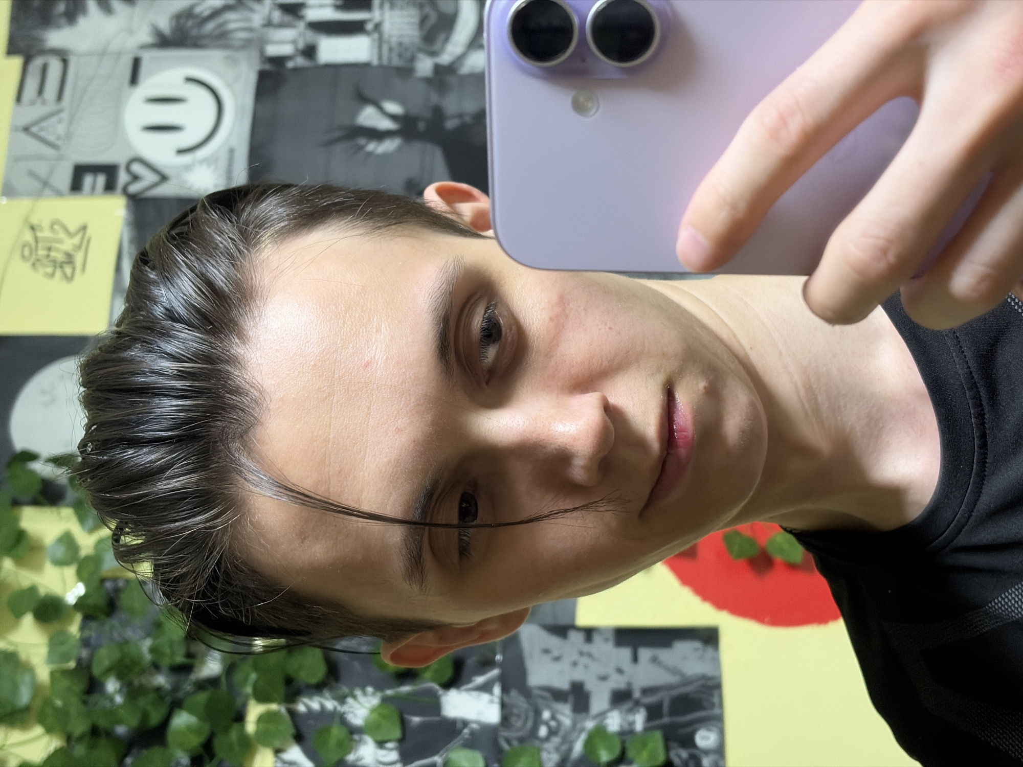 A young person is taking a selfie in a room, holding up a light purple smartphone. The person has dark, slicked-back hair with a prominent strand falling across their forehead. They are looking towards the phone camera with a slight smirk. The background consists of a wall covered in various posters and some artificial green foliage. One poster shows a smiley face, while others are black and white or yellow with abstract designs. The person is wearing a black t-shirt. The image appears to have been taken indoors, with artificial lighting. The location is unknown.