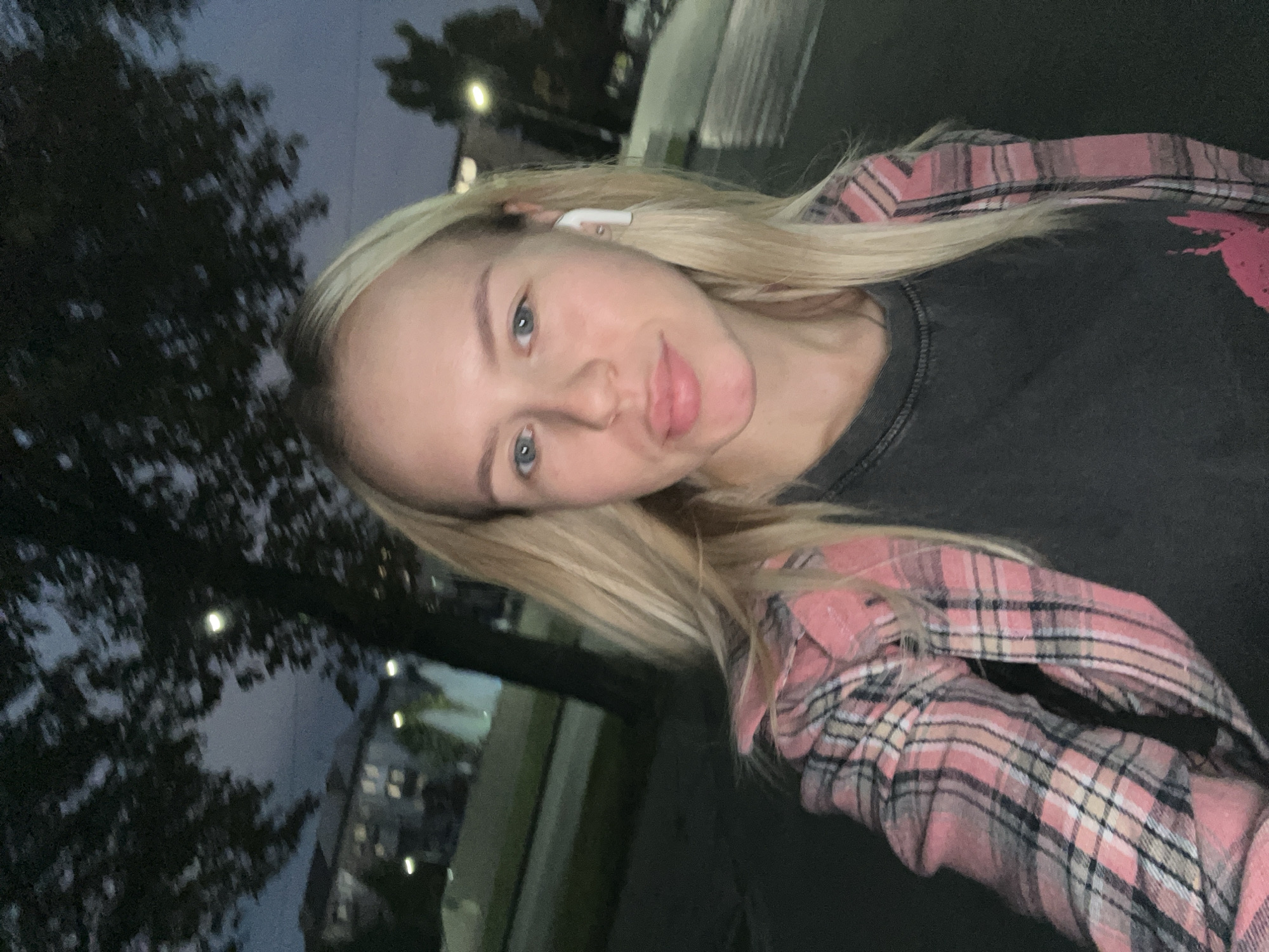 A young woman with blonde hair and blue eyes is captured in a close-up, outdoor selfie at dusk. She is wearing a dark t-shirt with a faded graphic and an open pink and gray plaid shirt. A white wireless earbud is visible in her right ear. The background suggests an urban setting with trees, buildings with illuminated windows, streetlights, and a wet street or sidewalk, indicating recent rain or damp conditions. The overall lighting is dim, characteristic of twilight.