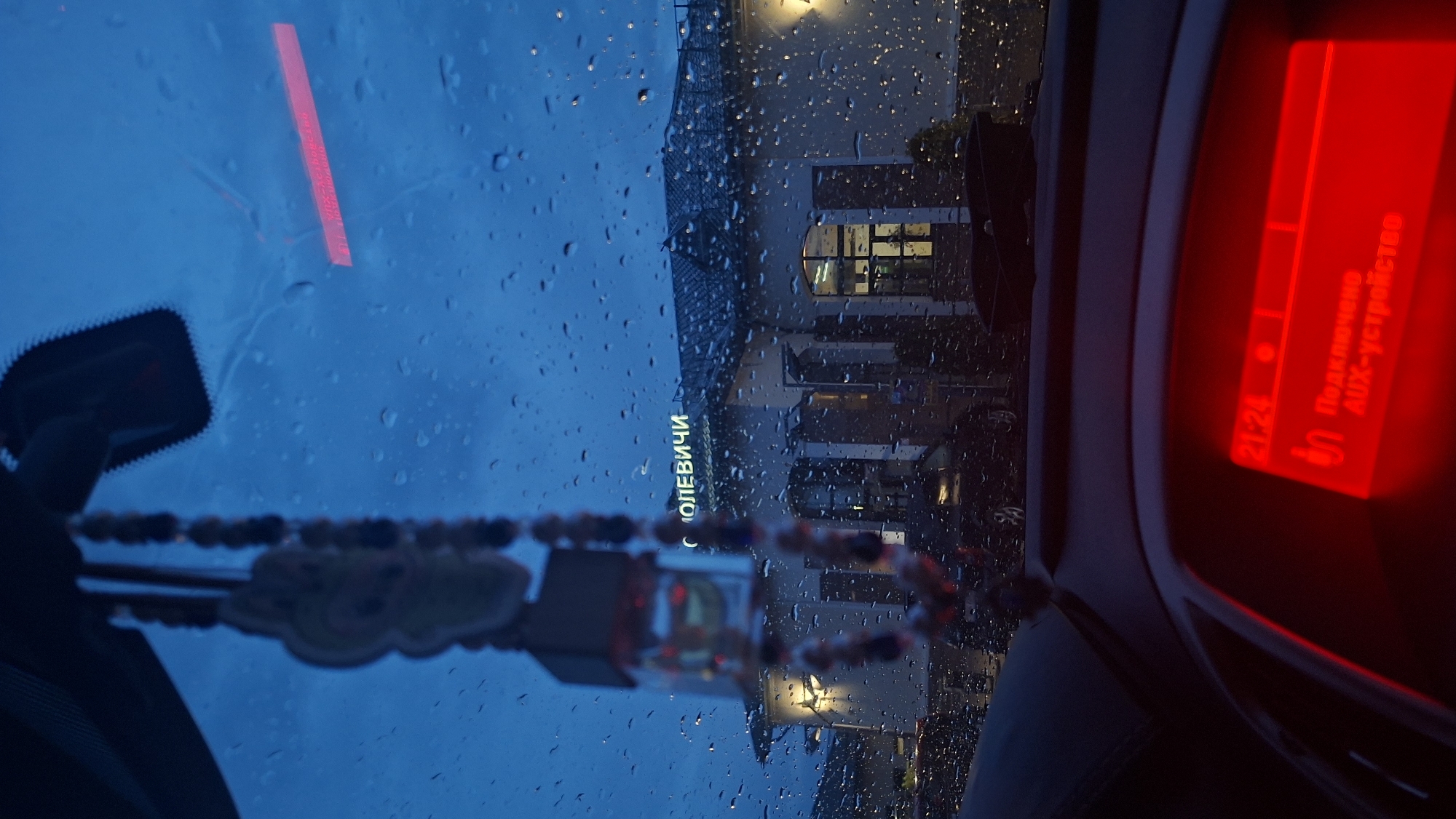 The photograph, taken from inside a vehicle on a rainy night, presents a blurred view of the street outside. Raindrops obscure the windshield. A red light, likely from the vehicle's dashboard, is visible on the right side of the frame. Hanging from the rearview mirror are decorative items. The building across the street has signage in a Cyrillic script, which appears to spell "С ОЛЕВИЧИ." The location is the city of Olevichi, Unknown.
