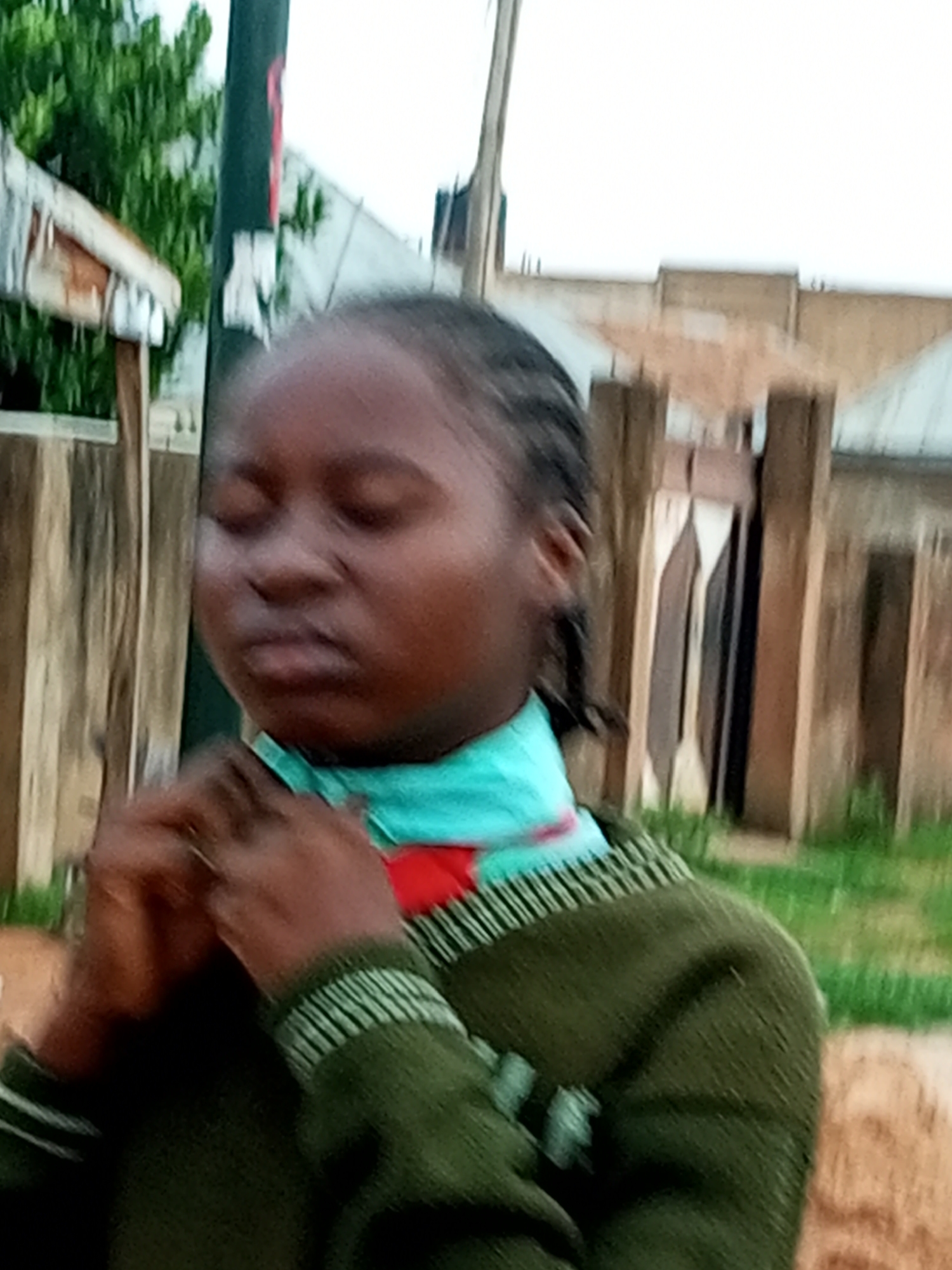 A young Black person with braided hair is shown from the chest up, facing forward with eyes closed and hands clasped in front of their chest. They are wearing a green sweater with a light blue and red scarf or high-necked garment underneath. The background is blurred, with elements suggesting an outdoor setting in a city environment, possibly Gombe, Nigeria. Visible in the background are wooden posts, a section of a building with a corrugated roof, and greenery. The overall image quality is low, with blurriness and pixelation, characteristic of a low-resolution photograph taken with a mobile device.