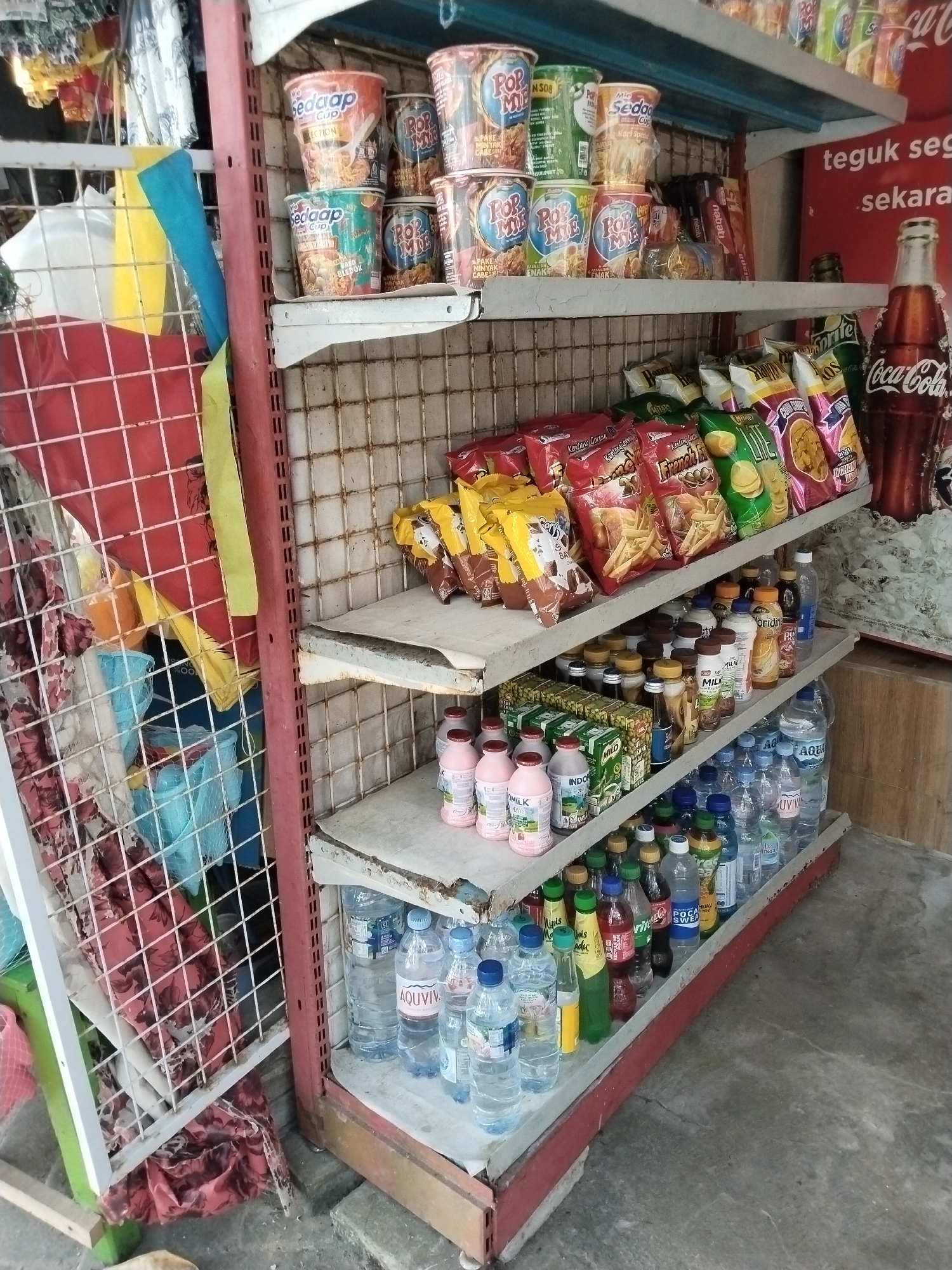 This is a photograph of a small convenience store or warung, likely located in Prigi, Indonesia. The scene depicts shelves stocked with various food and beverage items.
On the top shelf, there are several cups of instant noodles, including brands like "Sedap" and "Pop Mie." Bags of snacks, such as potato chips, are also visible. Further to the right, a large Coca-Cola bottle is prominently displayed, along with other bottles that are less distinct.
The middle shelf holds a variety of bottled drinks. There are bottles of what appear to be milk, juices, and water. Some smaller bottles, possibly condiments or sauces, are also present.
The bottom shelf is filled with more bottled water, including "AQUAVIVID" and "AQUA" brands. There are also numerous other beverage bottles, some appearing to be soft drinks.
In the foreground, on the left side of the image, a red wire rack is visible, containing some fabric items and plastic containers. The floor appears to be concrete. The overall environment suggests a modest, local retail establishment. There are no people visible in the frame.