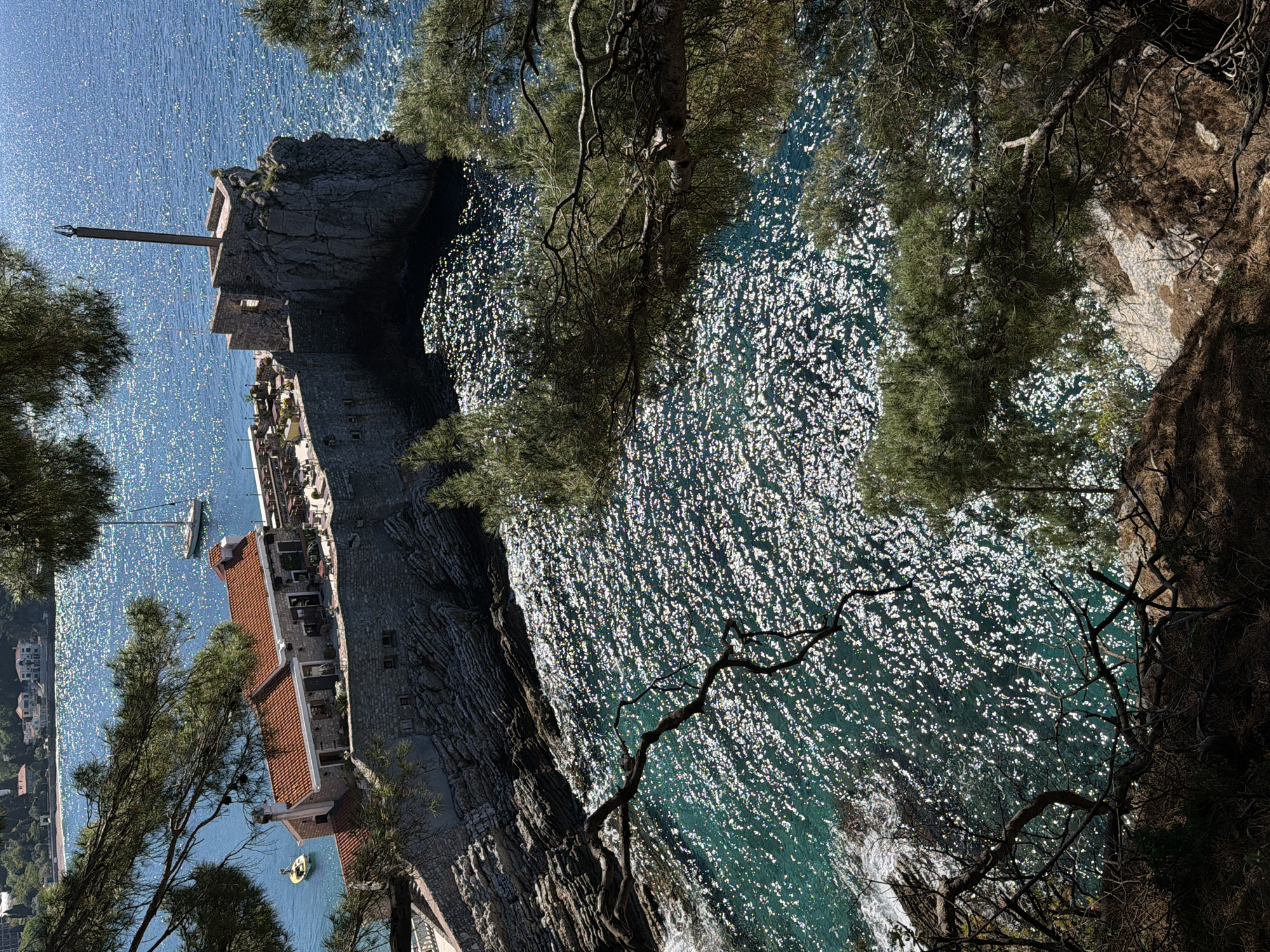 The image captures a vibrant coastal scene in Petrovac, Montenegro, viewed from a high vantage point looking down towards a rocky coastline and the sparkling Adriatic Sea. The orientation is rotated, but viewing it correctly, the scene unfolds under a bright, clear midday sun, indicated by the intense glitter of light reflecting off the blue-green water.
A cluster of stone buildings with distinctive red-tiled roofs is nestled into the rocky shore. One prominent structure features a tall, slender mast or pole extending upwards from a stone base, possibly a historical marker or navigational aid. Adjacent to this, other buildings appear to house amenities such as a restaurant or cafe, with an outdoor terrace visible along the waterfront.
The sea is a central element, its surface shimmering with countless points of light. Further out, a white sailboat is moored in the calm water, while closer to the shore, a small yellow inflatable boat with a person inside gently floats near the buildings. Two small figures are also discernible on a rocky outcrop near the mast-like structure.
The foreground and sides of the image are framed by dense, green pine trees with gnarled branches, growing on the rugged, earthy cliffside from which the photograph was taken. The combination of ancient stone architecture, lush vegetation, and the brilliant, sun-drenched sea evokes a serene and picturesque Mediterranean ambiance. No discernible text is visible in the image.
