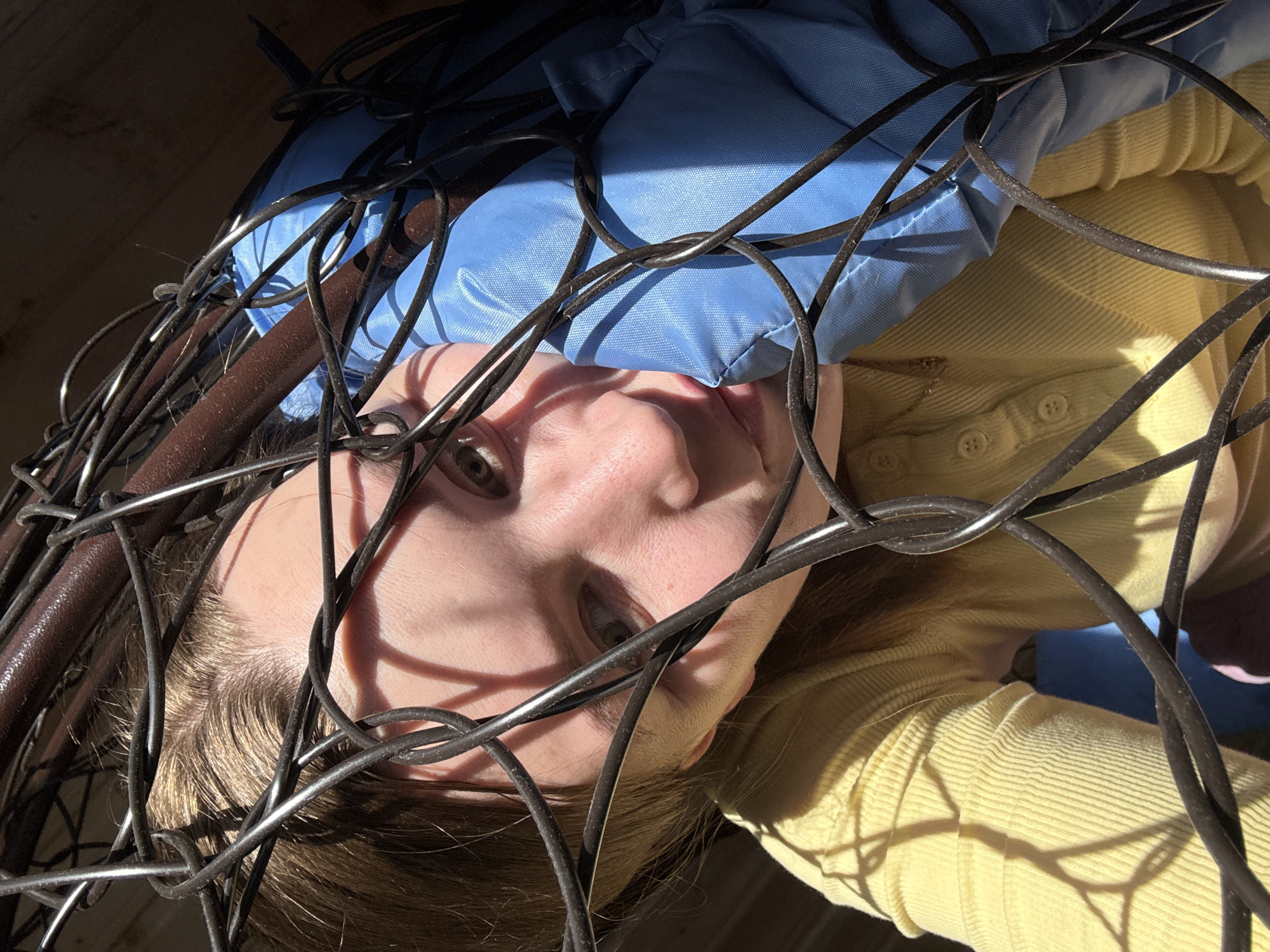 The scene is located in Italmas, Russia. A young individual, appearing female, with fair skin, light brown hair, and light-colored eyes, is positioned within or behind a complex structure of intertwined dark wires and brown tubular bars. The individual wears a light yellow, ribbed, long-sleeved top with a buttoned placket. To the right of the subject, a bright blue padded fabric object, possibly a cushion, is visible, partially entangled with the wire mesh. Strong, directional sunlight illuminates the scene from the right, casting sharp, elaborate shadows from the wire mesh onto the individual's face, hair, and clothing, creating a distinctive pattern. The individual maintains direct eye contact with the viewer, exhibiting a neutral expression. The background behind the structure is indistinct and dark.