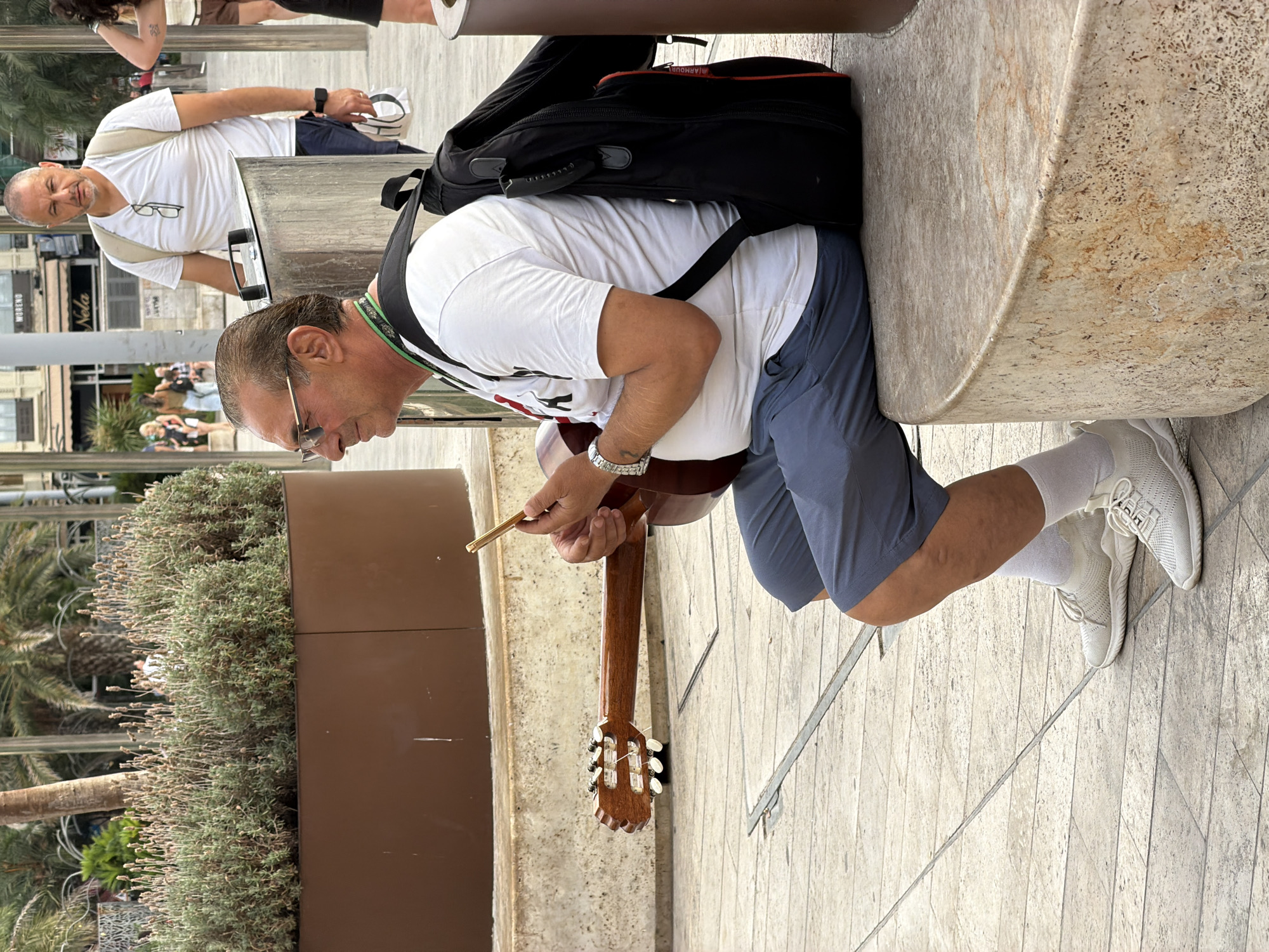 In an outdoor urban setting, a man in his 60s with tanned skin and graying hair sits on a curved, light-colored stone bench. He wears a white t-shirt, blue shorts, white socks, and white athletic shoes. He has on glasses with gold frames and a watch on his left wrist. He is holding a guitar, with the neck extending to his left, and appears to be looking at his smartphone. He is wearing a black backpack. Another man, appearing to be in his 50s, stands in the background to the left, wearing a white t-shirt, dark shorts, and a light-colored backpack, holding a white bag in his right hand. The background shows more people walking, buildings, palm trees, and a planter filled with lavender. The ground is paved with light-colored tiles.