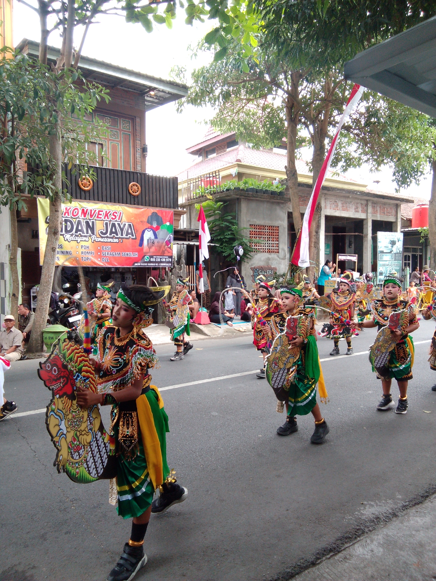 A group of children in traditional costumes, possibly a parade or performance, are walking down a street in Trenggalek, Indonesia. The children are adorned in elaborate, colorful outfits with headdresses and carry decorative props. One child in the foreground holds a carved wooden puppet-like object. Buildings line the street, including one with a banner advertising "Konveksi Dan Jaya" and various services like tailoring and school uniforms. A red and white Indonesian flag is visible, suggesting a national holiday or celebration. Spectators are seated and standing along the roadside, observing the procession. The environment appears to be an urban street with a paved road and sidewalks.