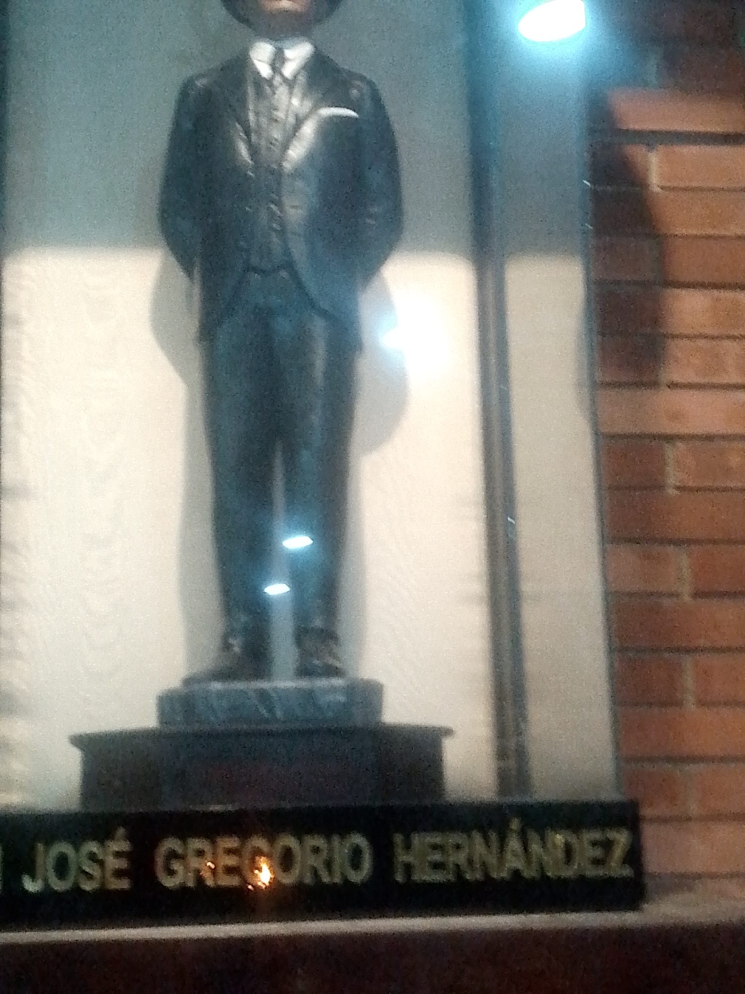 The image shows a statue of José Gregorio Hernández on display behind glass. The statue is dark in color, depicting a man in a suit, and is placed on a pedestal. A plaque bearing his name in gold lettering is visible at the base of the display. To the right of the glass display is a section of brick wall. The statue is a tribute to José Gregorio Hernández, a Venezuelan physician known for his piety and service to the poor. This statue is located in Caracas, Venezuela.