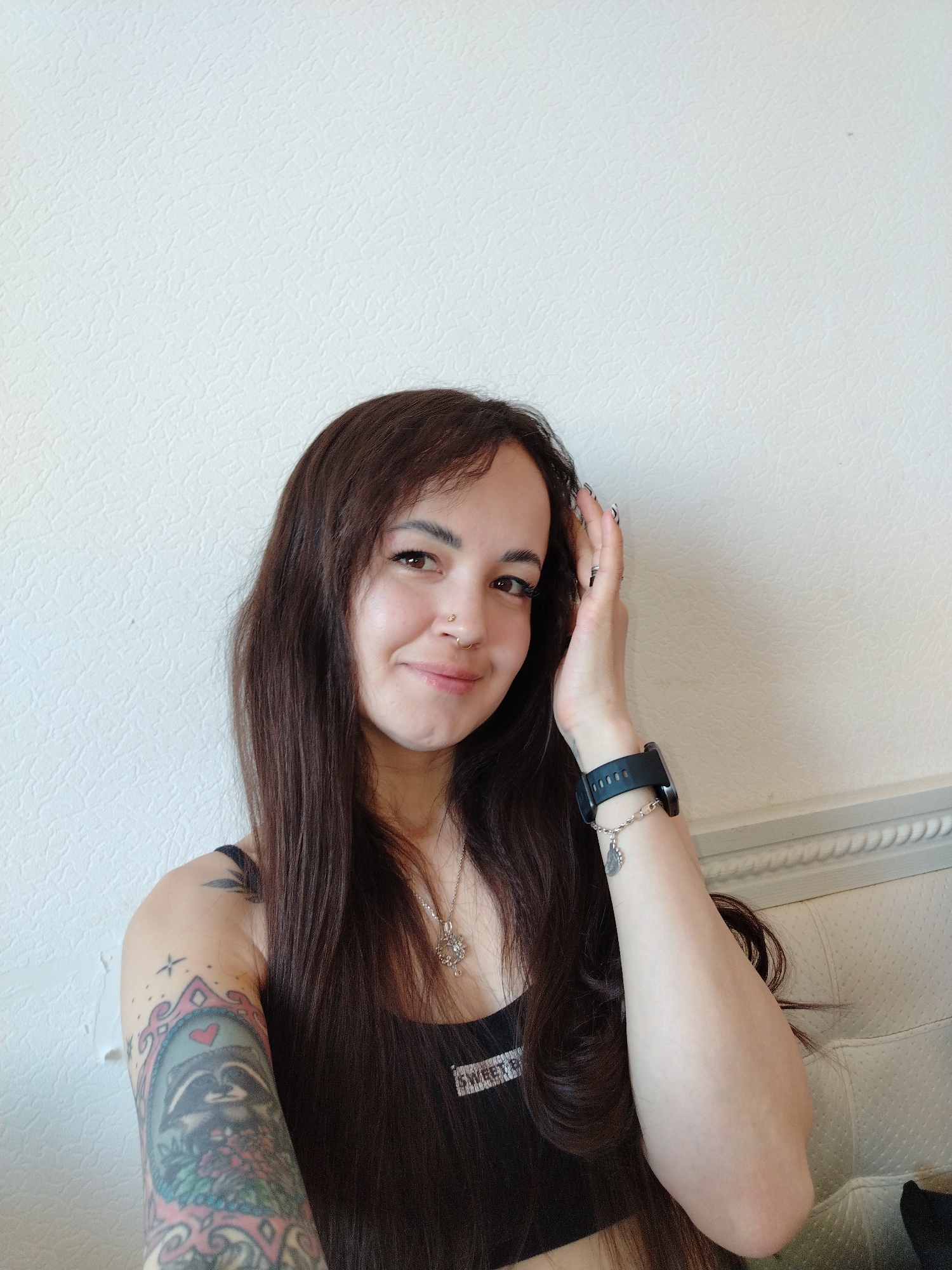 A young woman poses for a selfie indoors against a white wall with a textured surface. She is fair-skinned with long, dark brown hair. She is wearing a black tank top, a silver necklace, and a black smartwatch with a silver charm bracelet. She has a nose piercing on the left side. Her left arm is visible and displays a tattoo featuring a raccoon. She is smiling and holding her right hand up near her face, slightly touching her hair. The location is unknown, and there is no information to confirm if she is in Noviy, Russia.