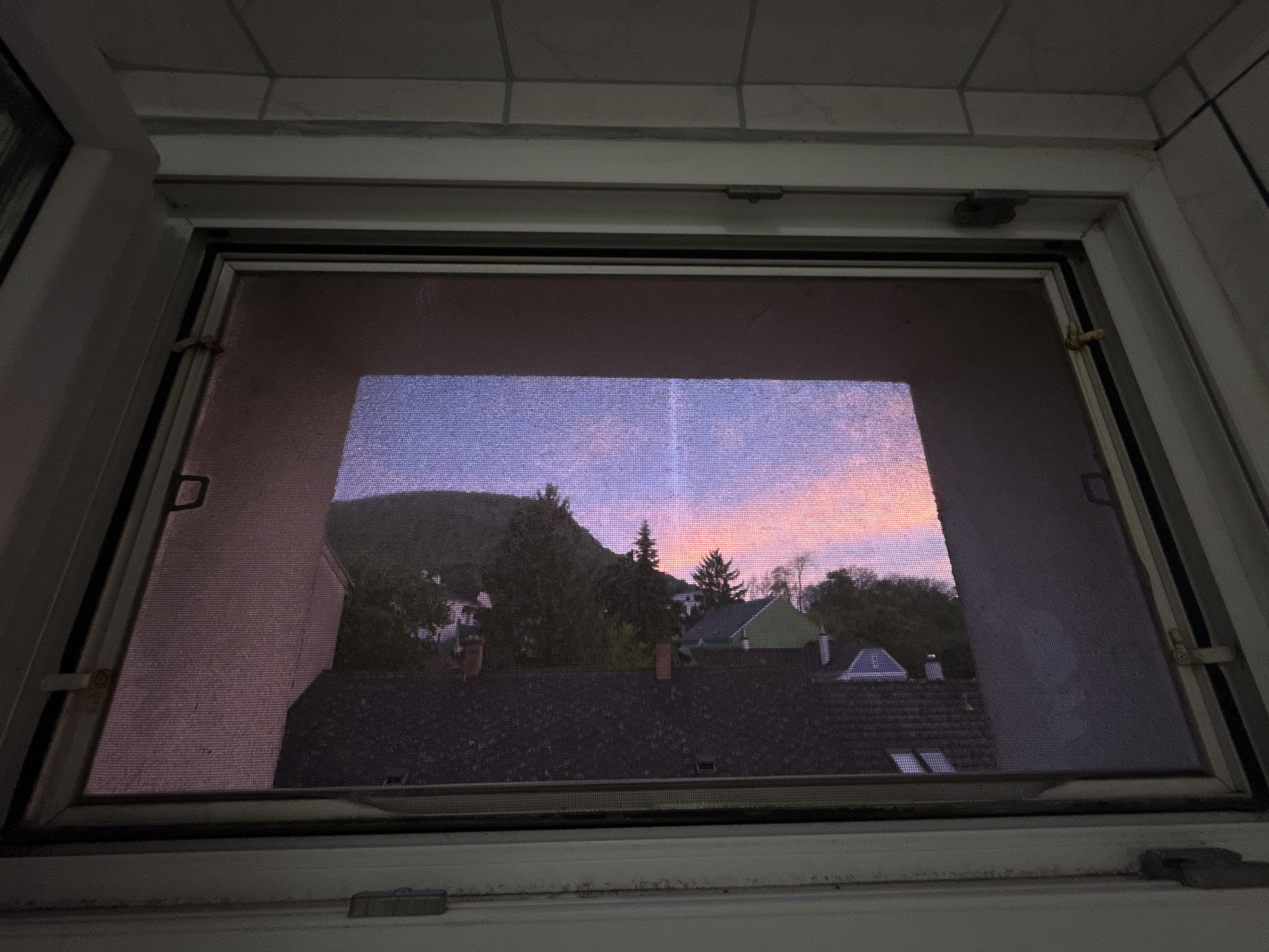 A view from inside a building, through a window with a screen, shows the town of Hainburg an der Donau, Austria, at dusk. The foreground is a window frame with a screen partially obscuring the view. Beyond the screen, rooftops of houses with varied shapes and colors are visible. Further in the background is a hillside with trees. The sky is a gradient of colors, transitioning from blue to pink as the sun sets. The image is taken at an oblique angle, looking slightly up and out. The lighting is dim, suggestive of late evening.