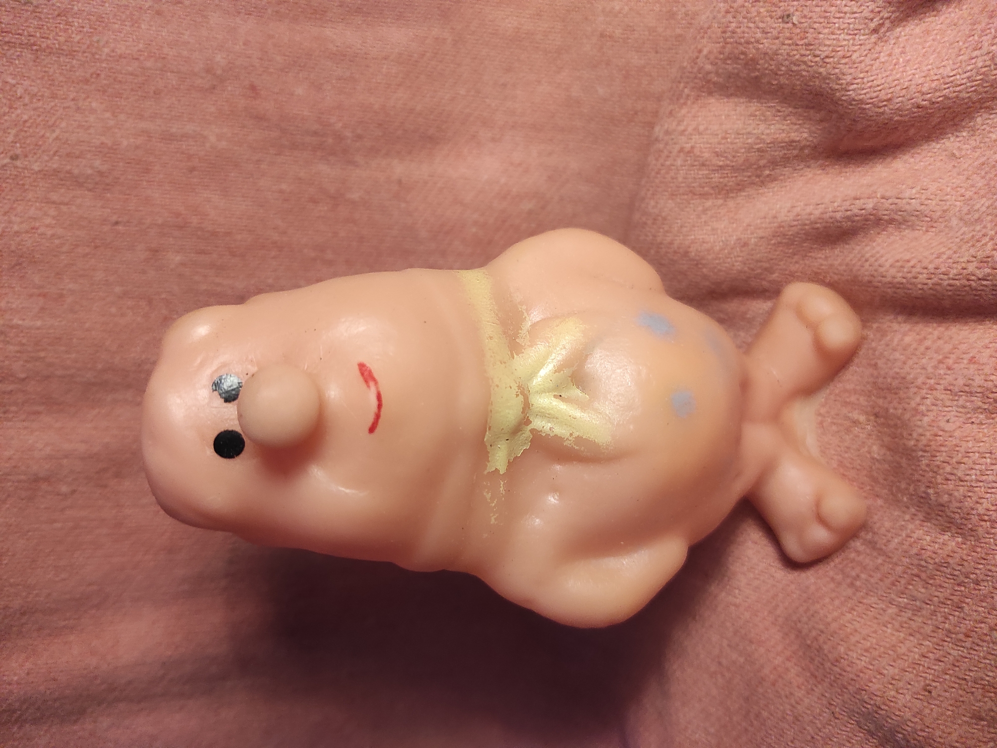 A pink, soft plastic toy figure stands upright on a textured pink fabric surface. The figure has a rounded head with two black dots for eyes, a small pink nose, and a red, curved mouth. A yellow scarf is tied around its neck. The body is bulbous, with stubby arms at its sides and two small feet at the base. Two light blue dots are painted on the front of the lower body. The fabric background is wrinkled and has a rough texture. The lighting appears to be from the left, casting a shadow to the right of the figure. The image was taken indoors, and no specific location in Chernihiv, Ukraine, can be identified from the visual information provided.