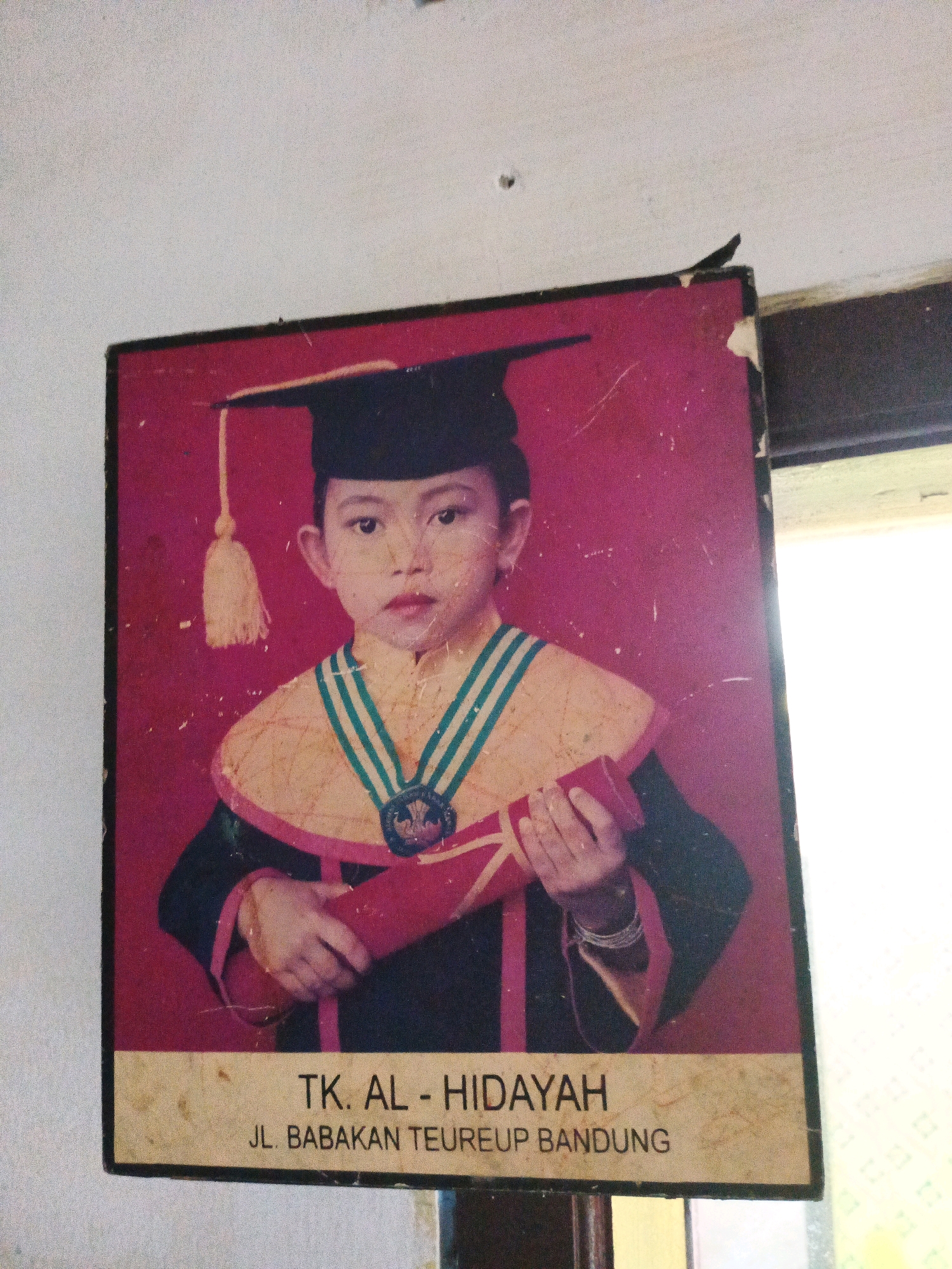 A framed photograph of a young child in a graduation cap and gown is displayed on a textured white wall, slightly askew. The child is holding a diploma, their gaze directed forward. The photograph is bordered with a tan strip containing text. The text at the top reads "TK. AL - HIDAYAH" and below it, "JL. BABAKAN TEUREUP BANDUNG". The photograph is slightly faded and scratched. A bright light source is visible to the right of the frame, suggesting natural light from a window or doorway. The overall environment appears to be indoors.