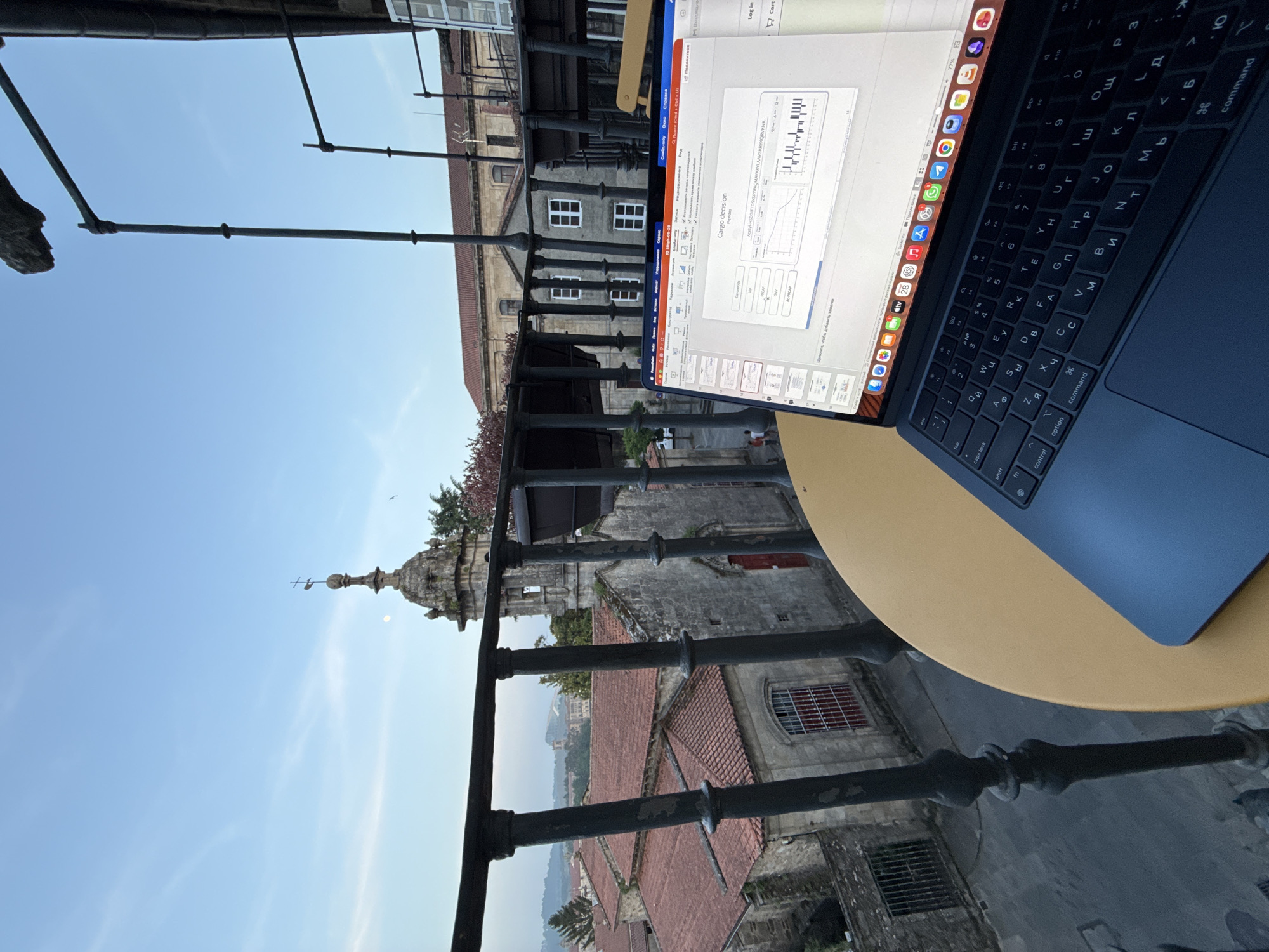 A laptop rests on a round table on a balcony, overlooking a cityscape in Santiago de Compostela, Spain. The keyboard layout appears to be Cyrillic. The laptop screen displays a presentation slide, possibly related to research or business analytics. The balcony has a wrought iron railing, behind which are buildings with red-tiled roofs, stone facades, and architectural details indicating a historic area. The sky is clear and blue, suggesting daylight. A spire or tower can be seen in the background, possibly part of a cathedral or significant historical structure.