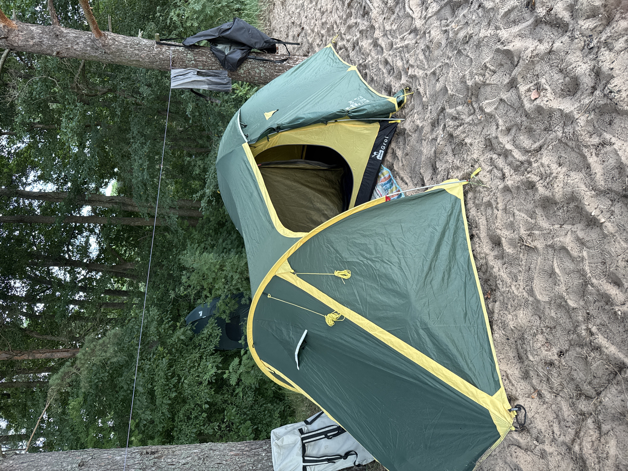 In the city of Zelenaya Roshcha, Russia, a campsite is set up on a sandy area near a forest. A green and yellow tent is pitched in the foreground, its entrance open, revealing the yellow interior. To the left of the tent, a light-colored backpack with black straps rests on the sand. A clothesline stretches between two trees, with clothing and a black backpack hanging from it. The background is dominated by a dense forest of green trees. The sandy ground is marked with numerous footprints.
