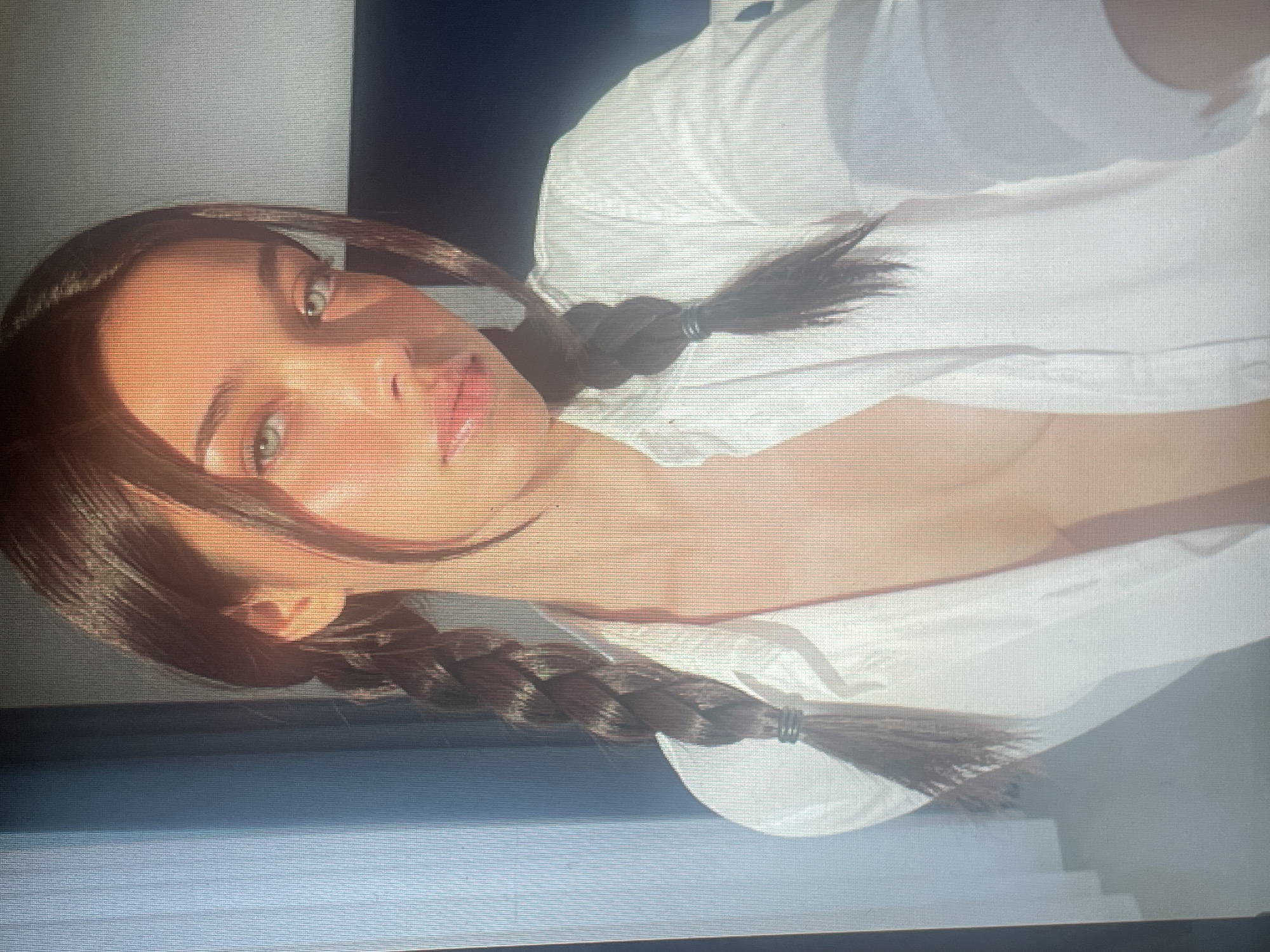 The image is a medium close-up of a young woman with dark brown hair styled in two braids. She is wearing an open white shirt, revealing her chest. Her eyes are a light green color, and she has a neutral expression. The lighting suggests it is daytime, with sunlight casting shadows on her face and shirt. The background is out of focus, appearing to be an indoor setting with some vertical blinds on the left and a dark surface on the right. There is no discernible information about the location being in Mantov, Czechia.