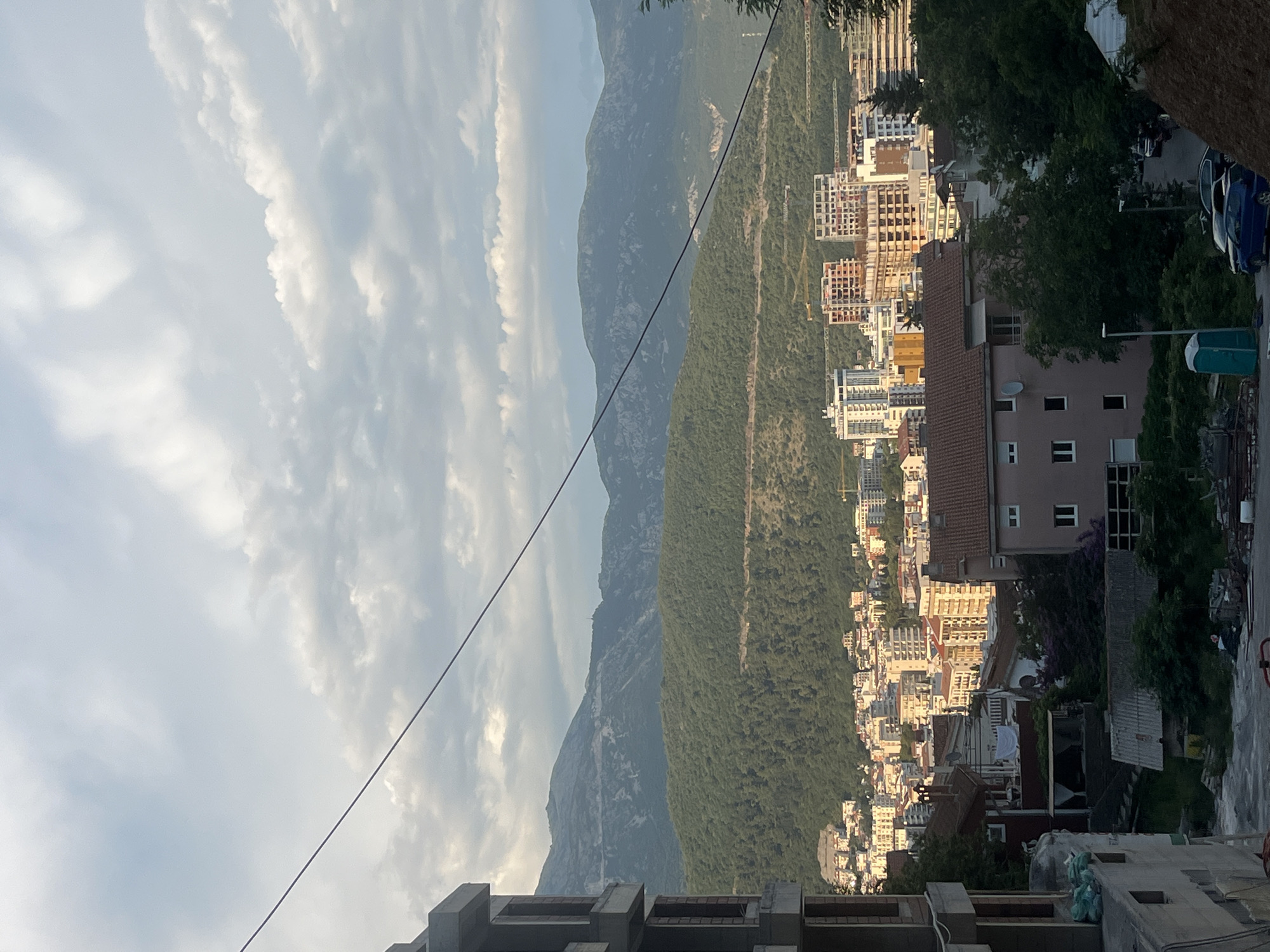 The image captures a view of Budva, Montenegro, characterized by a densely built urban landscape set against a backdrop of lush, green hills and distant, rugged mountains under a cloudy sky. In the foreground, an unfinished concrete building on the left suggests ongoing construction. Below this, a collection of residential buildings, some with red-tiled roofs, are clustered together, interspersed with trees. Further into the scene, a more modern part of the city is visible, featuring taller buildings, including some under construction, with cranes dotting the skyline. The middle ground is dominated by the verdant slopes of the hills, with winding roads visible on their inclines. The sky above is partly cloudy, with patches of blue peeking through. A thin black wire runs diagonally across the upper portion of the image. The overall impression is one of a coastal city expanding into its natural surroundings.