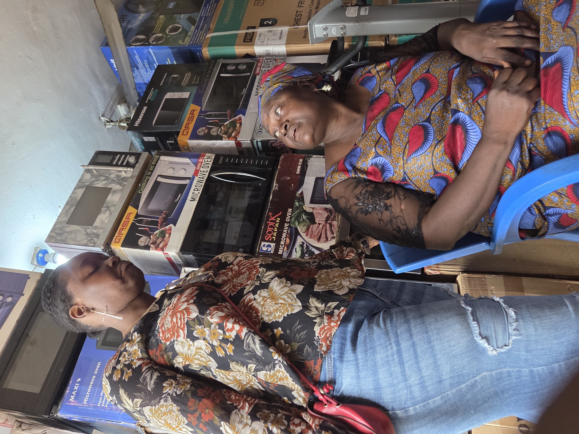 The image depicts two Nigerian women in an indoor setting, likely a store, characterized by stacks of boxed appliances, predominantly microwaves, in Jos, Nigeria. The woman on the left, facing away from the camera and slightly towards the right, wears a dark, long-sleeved shirt with a colorful floral print and light blue ripped jeans. She has dark hair braided and a cross-shaped earring in her left ear. She carries a red crossbody bag. The woman on the right is seated in a blue plastic chair, looking upwards and to her left. She wears a headscarf and a vibrant, patterned dress with blue, red, and yellow designs, and has lace detailing on her sleeves. Her hands are clasped in her lap. The background is filled with numerous boxes for electronic appliances, suggesting a retail environment for household electronics. The lighting appears to be artificial, coming from overhead.
