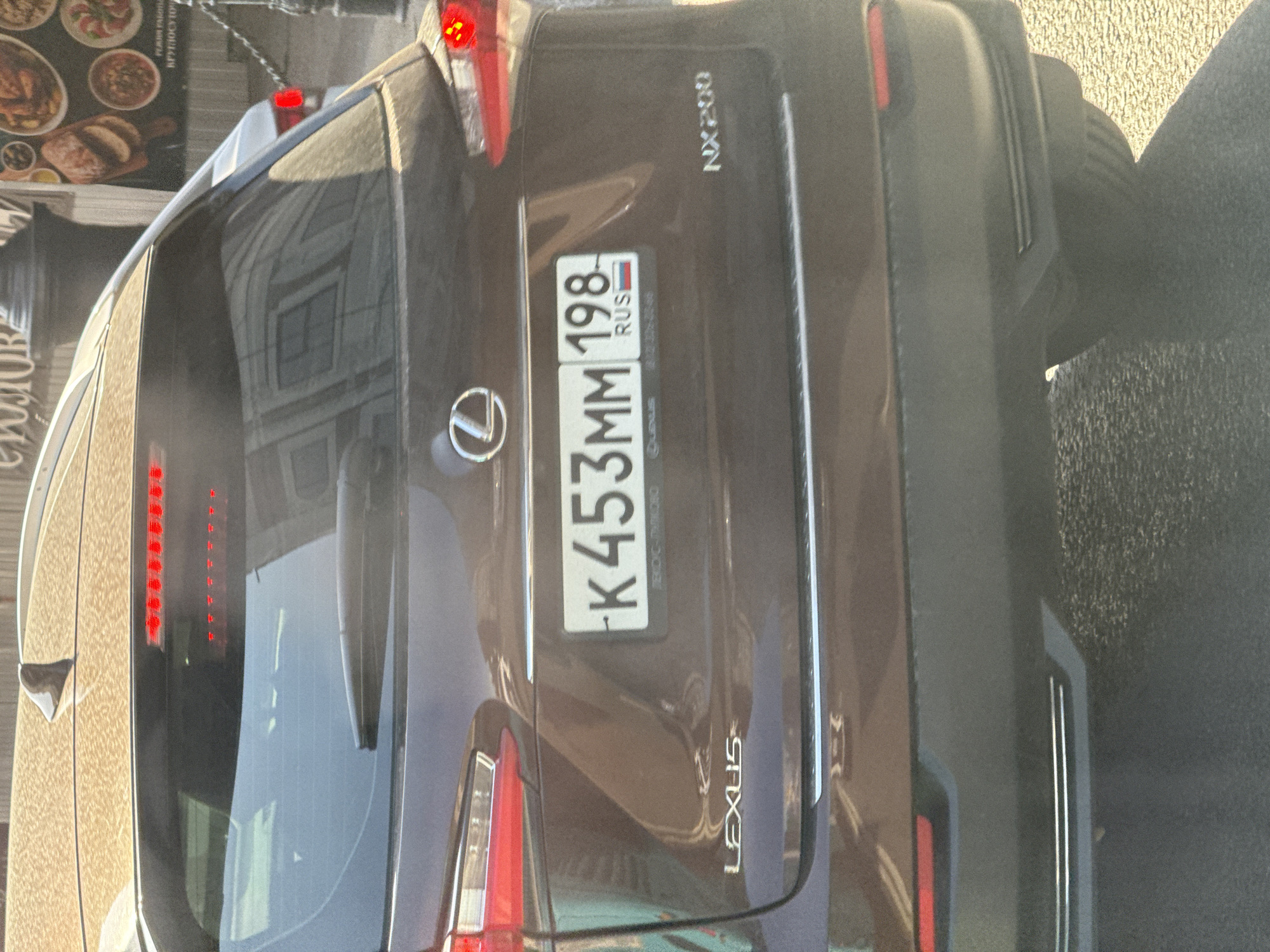 A dark brown Lexus NX200 is parked on a street in Saint Petersburg, Russia. The car's rear is facing the camera, and its license plate reads "K453MM 198 RUS." The car is parked on asphalt, and a building is visible in the background, partially obscured by reflections on the car's windows. The overall lighting suggests it is daytime.