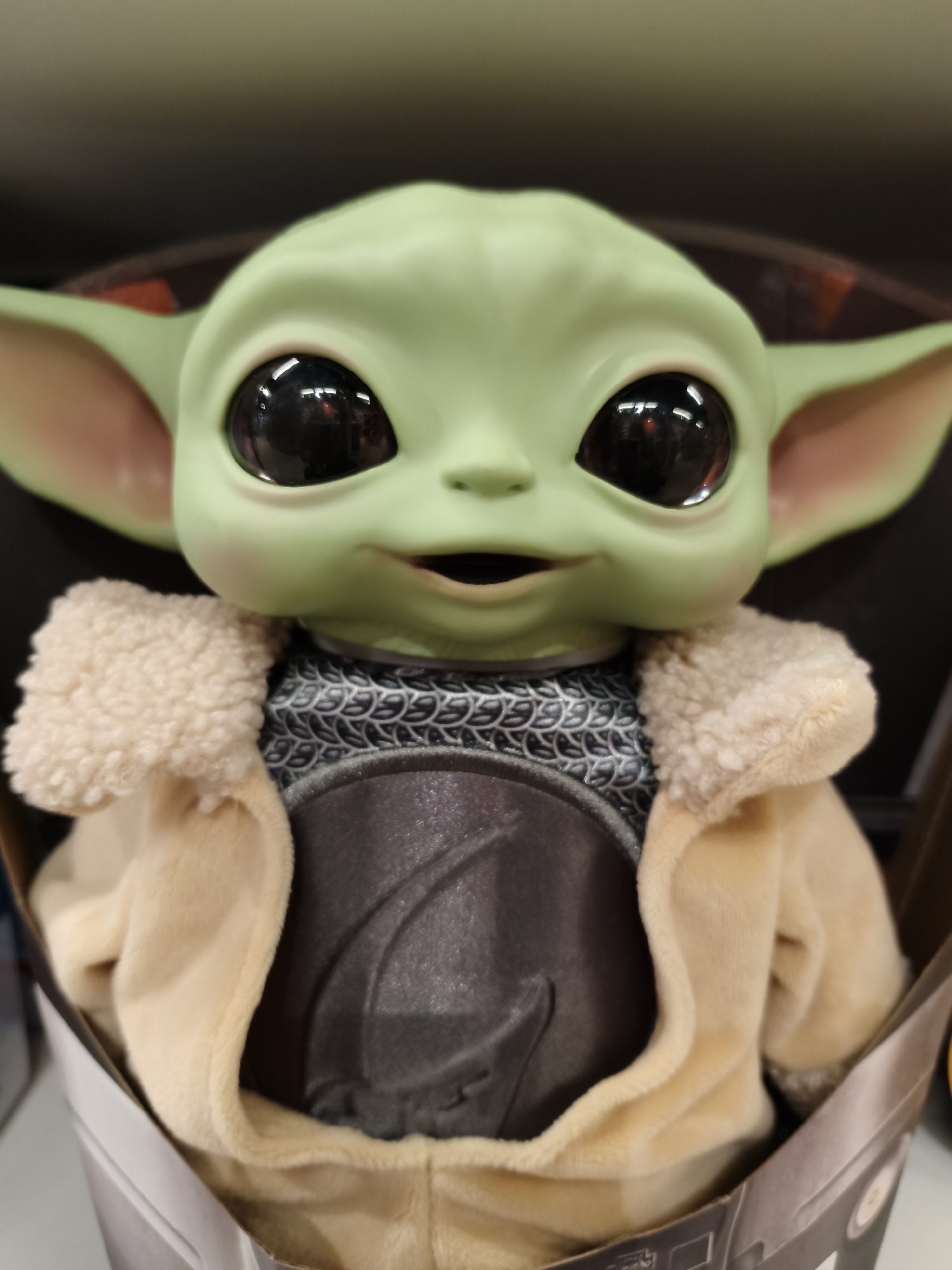 A close-up, eye-level shot captures a toy resembling Grogu (Baby Yoda) from Star Wars: The Mandalorian, presented in what appears to be its retail packaging. The toy, made of light green plastic or silicone, features large, shiny black eyes that reflect indistinct light sources and colors from the surrounding environment, suggesting an indoor setting. Its small mouth is slightly agape, giving it a neutral to mildly curious or happy expression.
The toy is dressed in a soft, light tan or cream-colored coat with a plush, sherpa-like collar, mimicking Grogu's signature attire. Beneath the coat, a darker, patterned garment resembling chainmail or scales is visible around the chest and neck area, along with a darker, smooth panel over the abdomen. The packaging, visible at the bottom and sides, is made of cardboard, with part of a dark grey and white design discernable. The background is dark and out of focus, further emphasizing the toy. No discernible text is visible on the toy or its immediate packaging. The image likely depicts a product display in a store or an unboxing scenario in Dnipro, Ukraine.